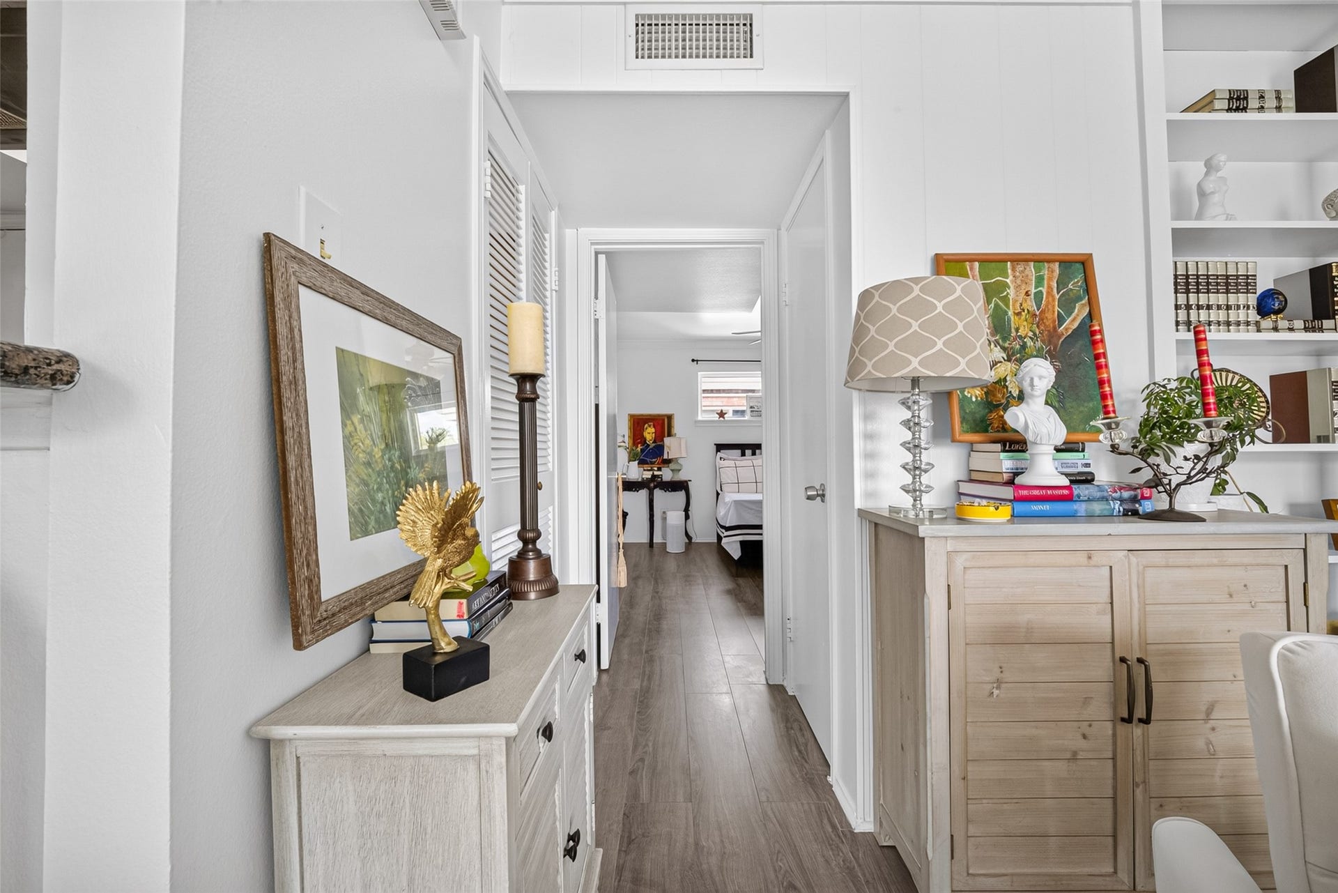 6402 Del Monte Drive, #65, Houston, TX 77057 This photo showcases a bright and inviting hallway leading to a bedroom. The space features elegant decor with bookshelves, framed art, and a stylish lamp on a wooden cabinet. The light wood flooring and white walls add to the airy feel.
