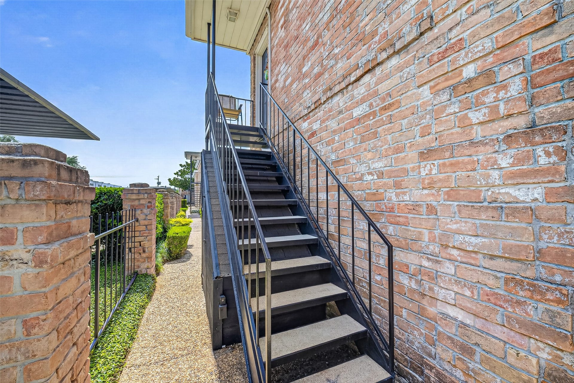 6402 Del Monte Drive, #65, Houston, TX 77057 This photo shows an exterior metal staircase attached to a brick wall, leading to an upper level. The path alongside is paved and bordered by well-maintained greenery and a wrought iron fence.