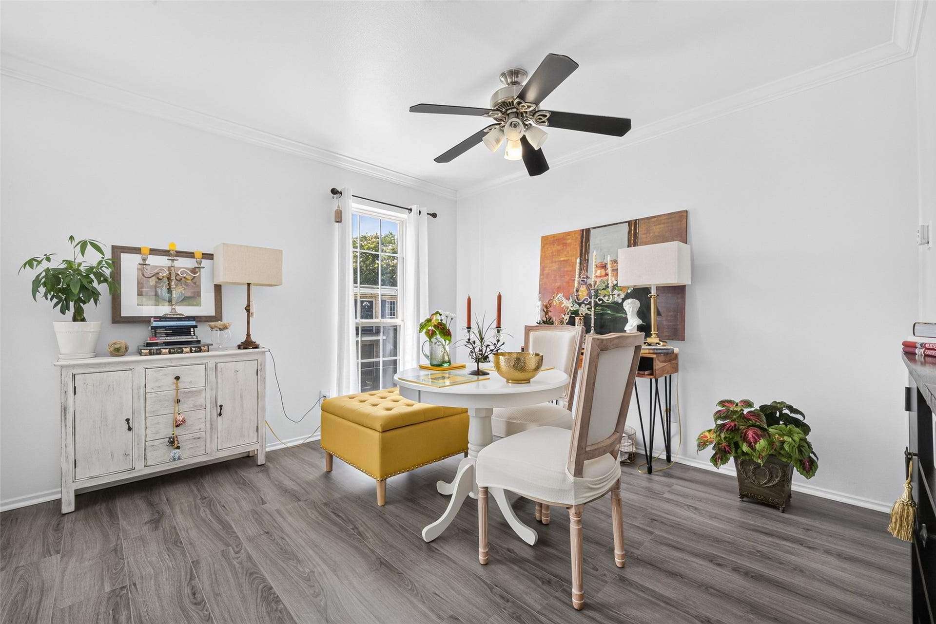 6402 Del Monte Drive, #65, Houston, TX 77057 This bright dining area features elegant decor with a round white table, cushioned chairs, and a stylish mustard ottoman. Warm wood flooring complements the white walls, while large windows and a ceiling fan enhance the airy feel. Decor includes plants, artwork, and tasteful lighting.