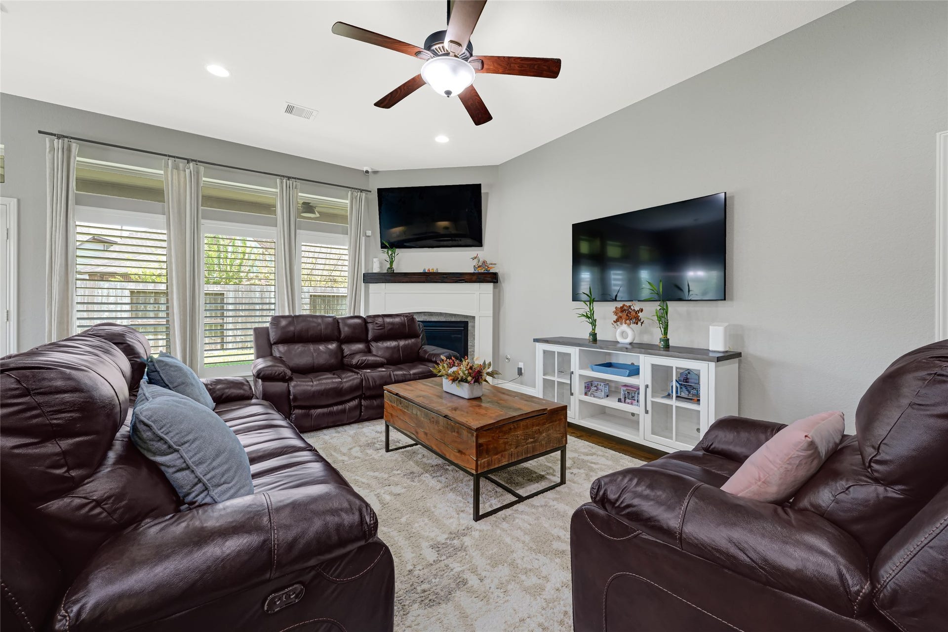 The inviting family room features a cozy fireplace, expansive backyard views through elegant plantation shutters with soft-colored curtains, and includes two TVs for your entertainment.