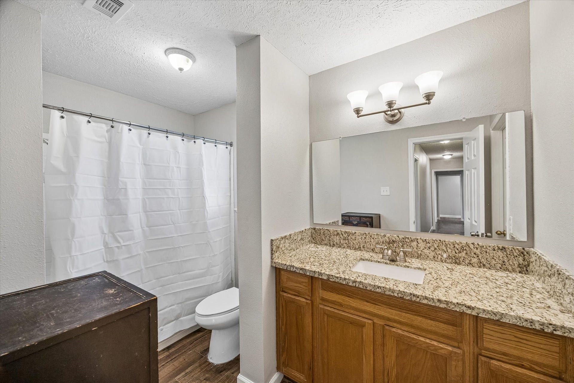 Bright bathroom featuring a spacious granite-topped vanity, a large mirror, and a shower, complemented by warm wood cabinetry and elegant lighting.