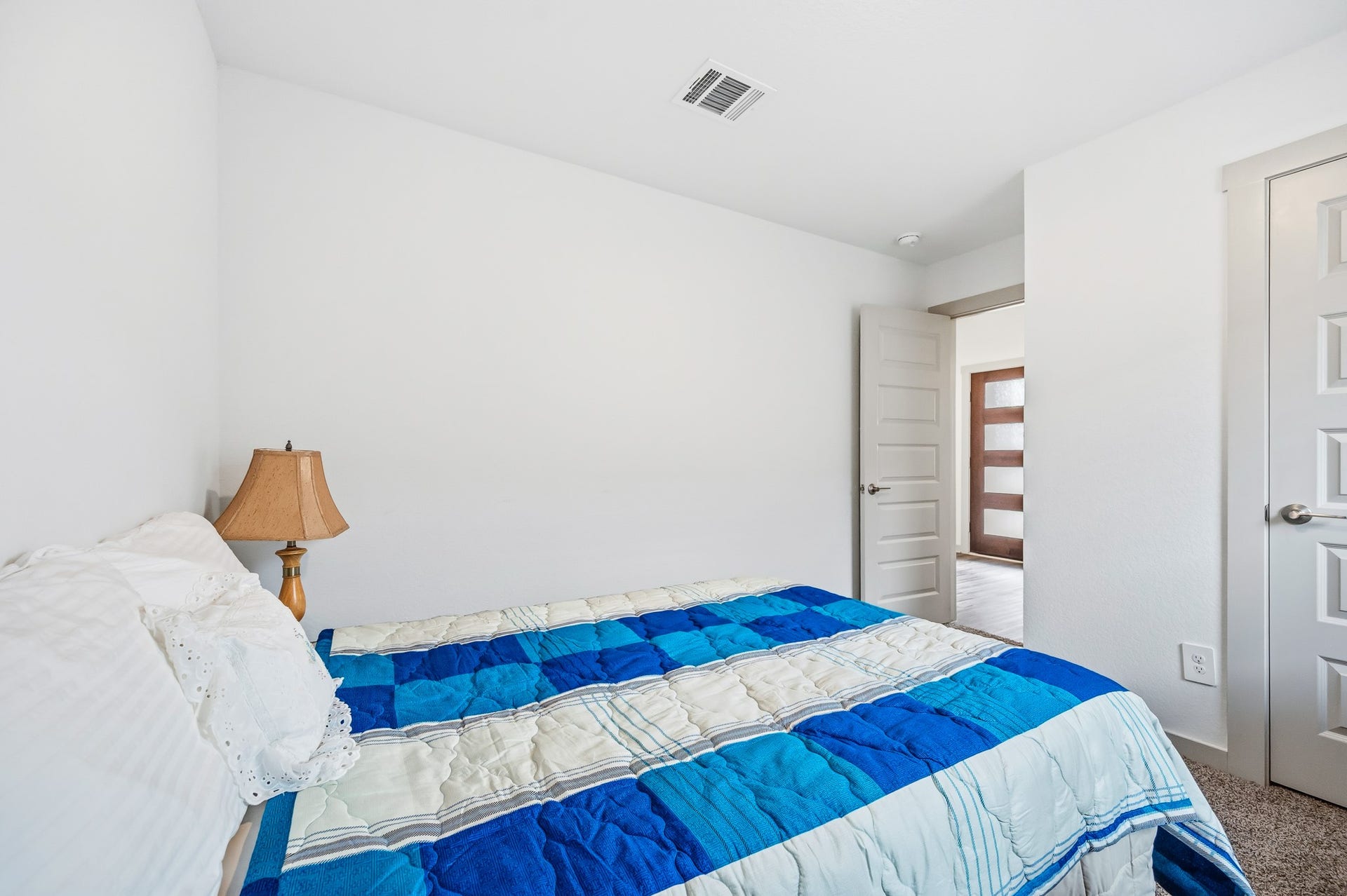 7002 Fleetwood Crescent Way, Richmond, TX 77407 This cozy bedroom features a modern design with white walls, a blue and white quilt on the bed, and a bedside lamp. The room is well-lit and has an open door leading to a hallway, enhancing the sense of space.