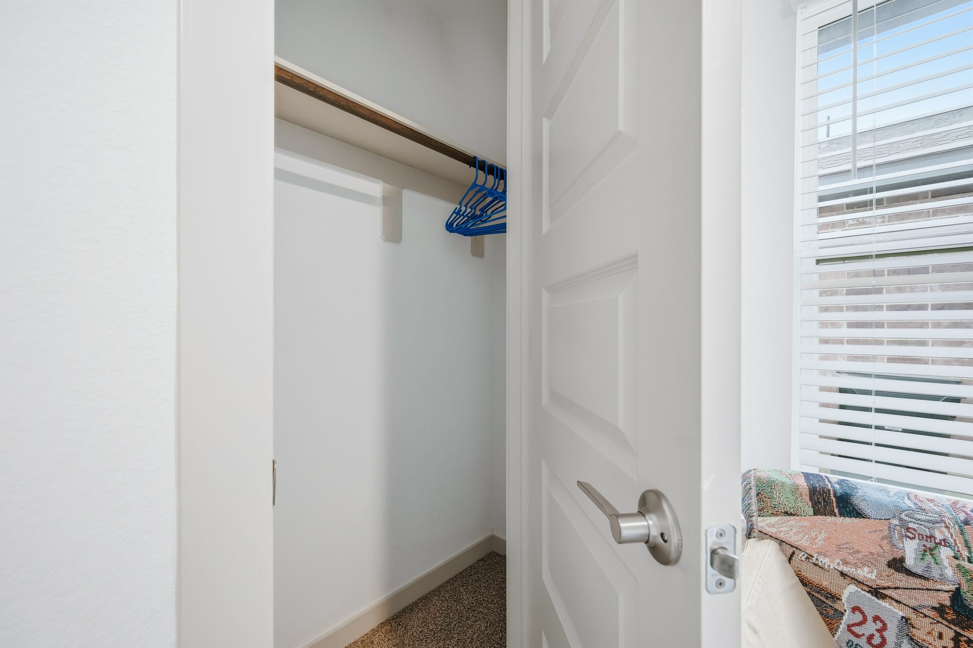 7002 Fleetwood Crescent Way, Richmond, TX 77407 A small, white closet with a wooden rod and blue hangers, carpeted flooring, and an open door. Nearby, there's a window with blinds allowing natural light to enter.