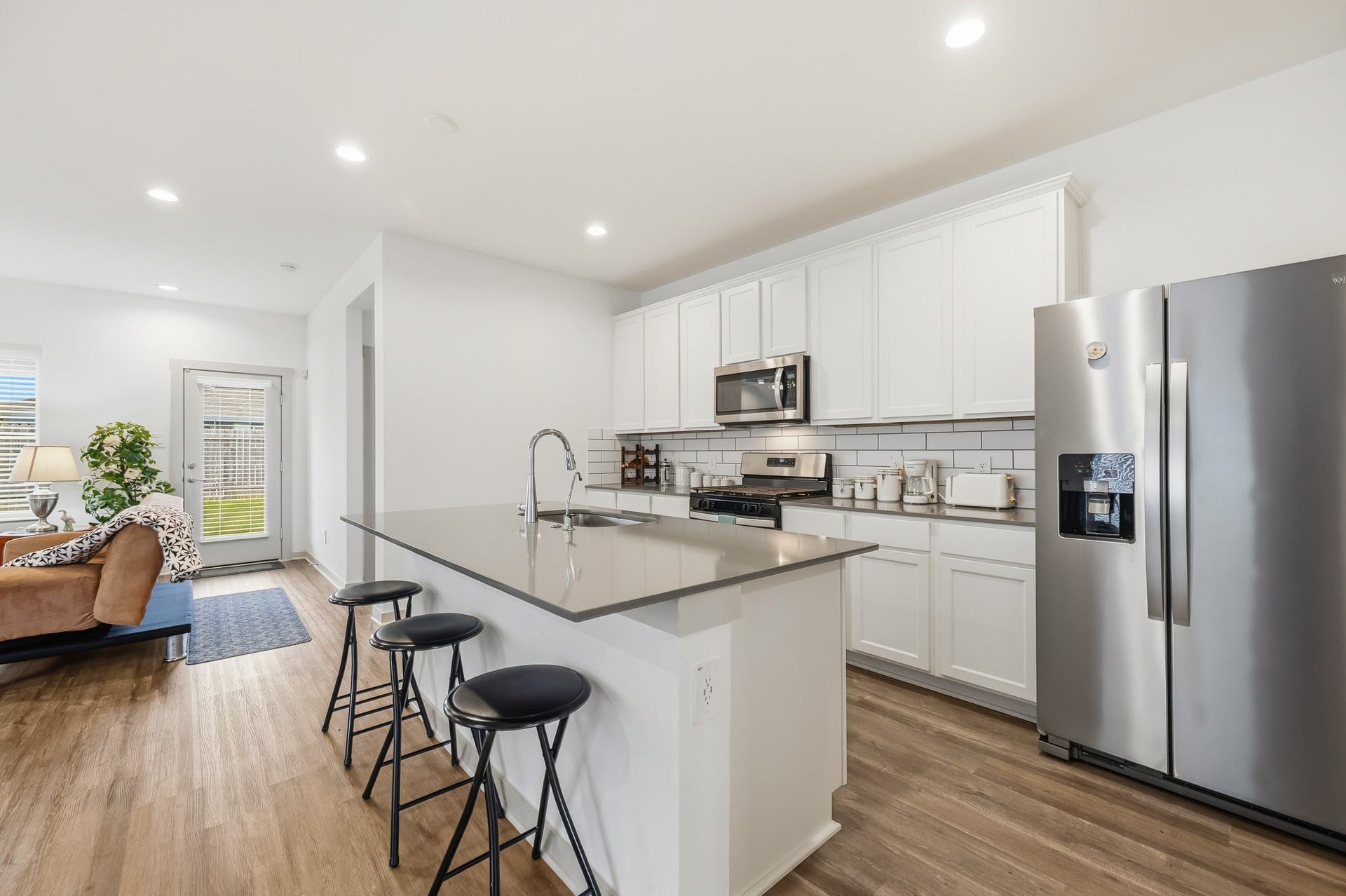 7002 Fleetwood Crescent Way, Richmond, TX 77407 This modern kitchen features sleek white cabinetry, stainless steel appliances, and a spacious island with bar seating. It opens into a cozy living area with hardwood floors and natural light from a glass door leading outside.