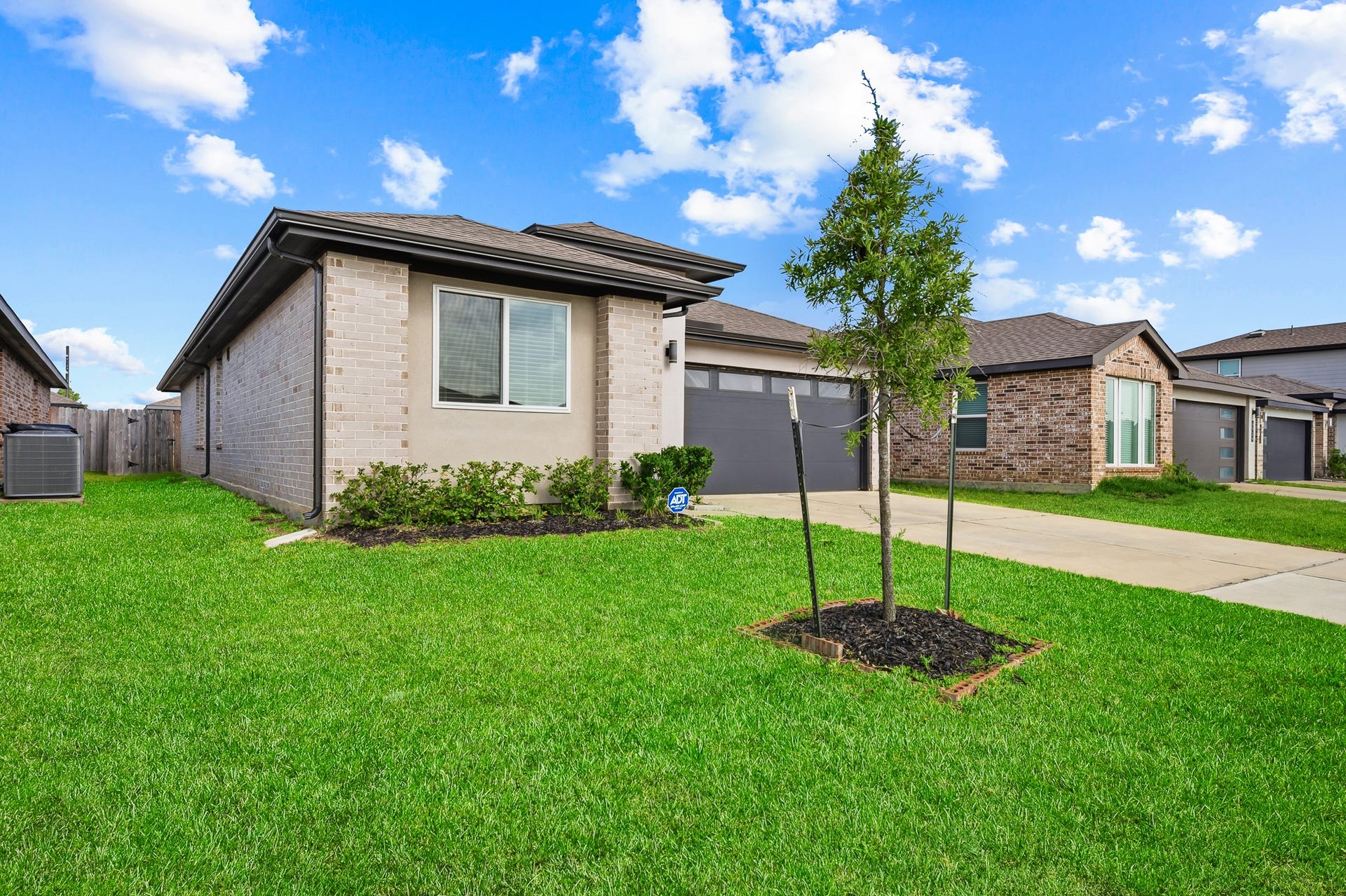 7002 Fleetwood Crescent Way, Richmond, TX 77407 This photo shows a modern, single-story brick home with a well-maintained lawn and a young tree in the front yard. The house has a light brick exterior, large windows, and a two-car garage. It's situated in a suburban neighborhood with similar style homes.