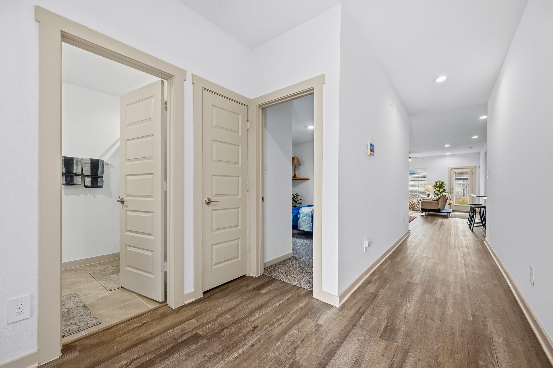 7002 Fleetwood Crescent Way, Richmond, TX 77407 This photo showcases a modern hallway with wood-style flooring, leading to a bright living area. It features a bathroom on the left and a bedroom on the right, both with beige door frames. The space is well-lit with recessed lighting.