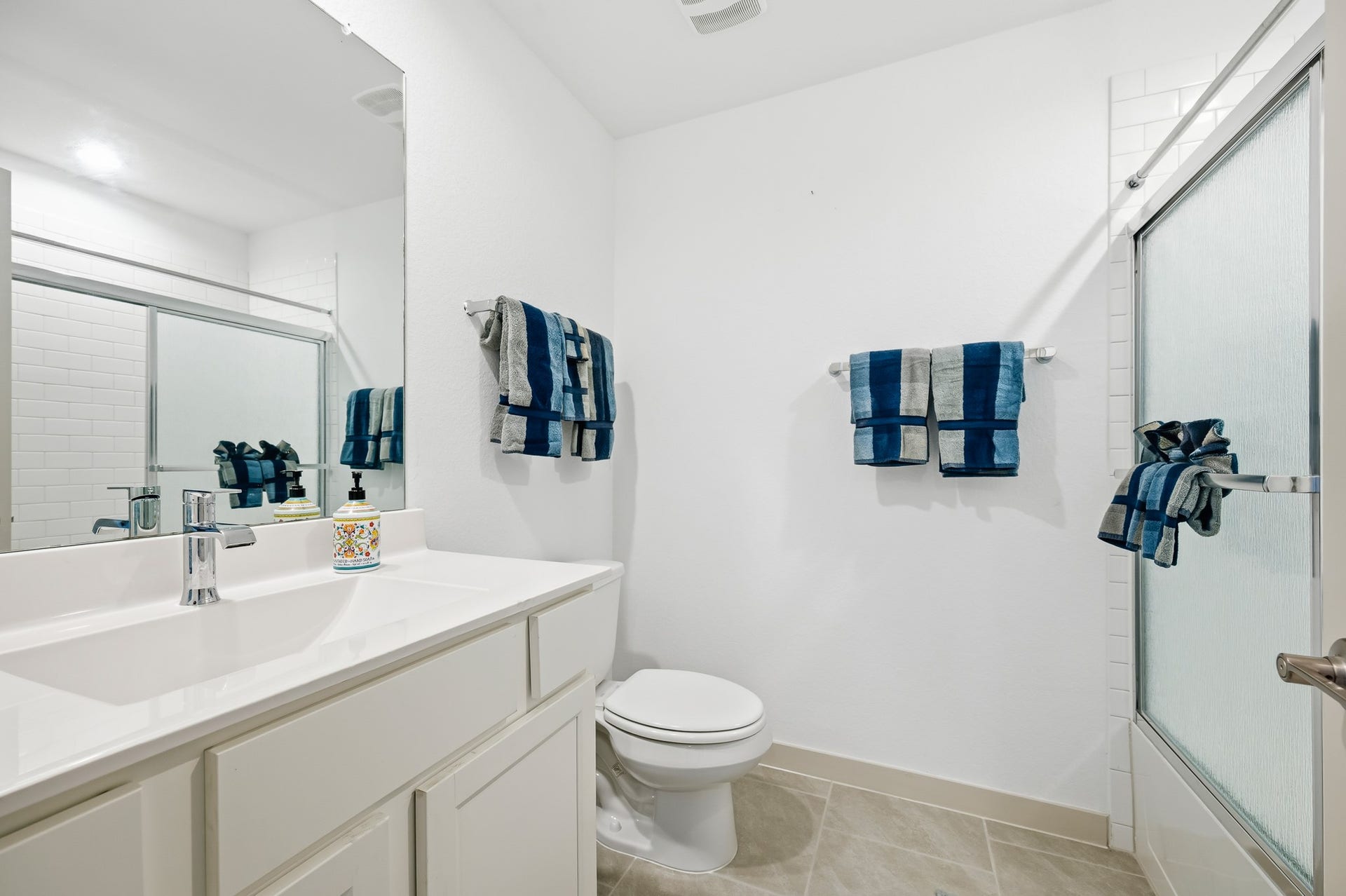 7002 Fleetwood Crescent Way, Richmond, TX 77407 This bathroom features a sleek design with a large mirror, a modern vanity, and a glass-enclosed shower. The space is accented with blue and gray towels, creating a clean and inviting atmosphere.