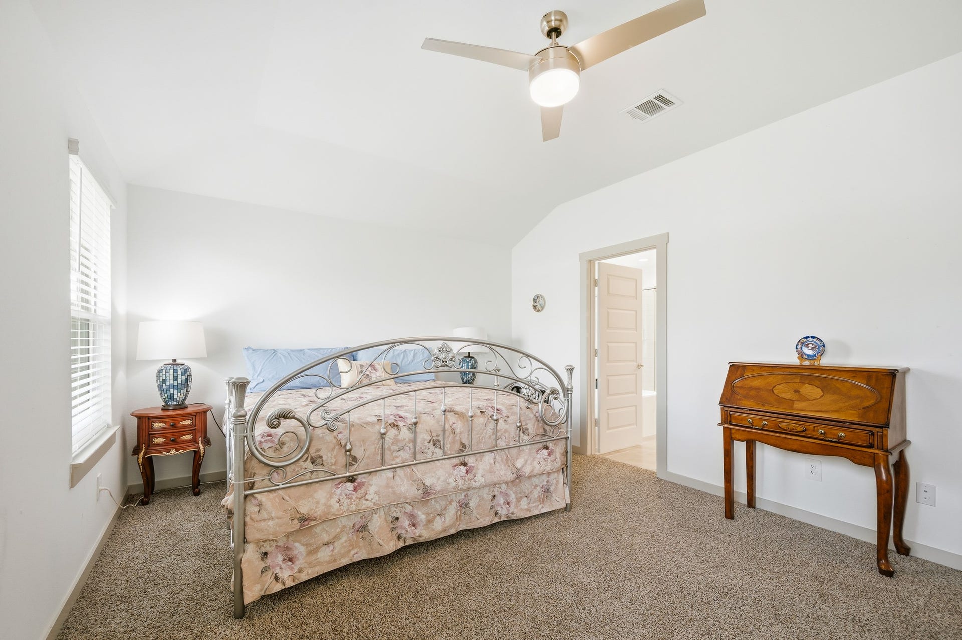 7002 Fleetwood Crescent Way, Richmond, TX 77407 This bedroom features a cozy setting with neutral walls and carpet, a decorative metal bedframe, and ample natural light from a large window. A ceiling fan adds comfort, while a wooden side table and desk offer classic charm.