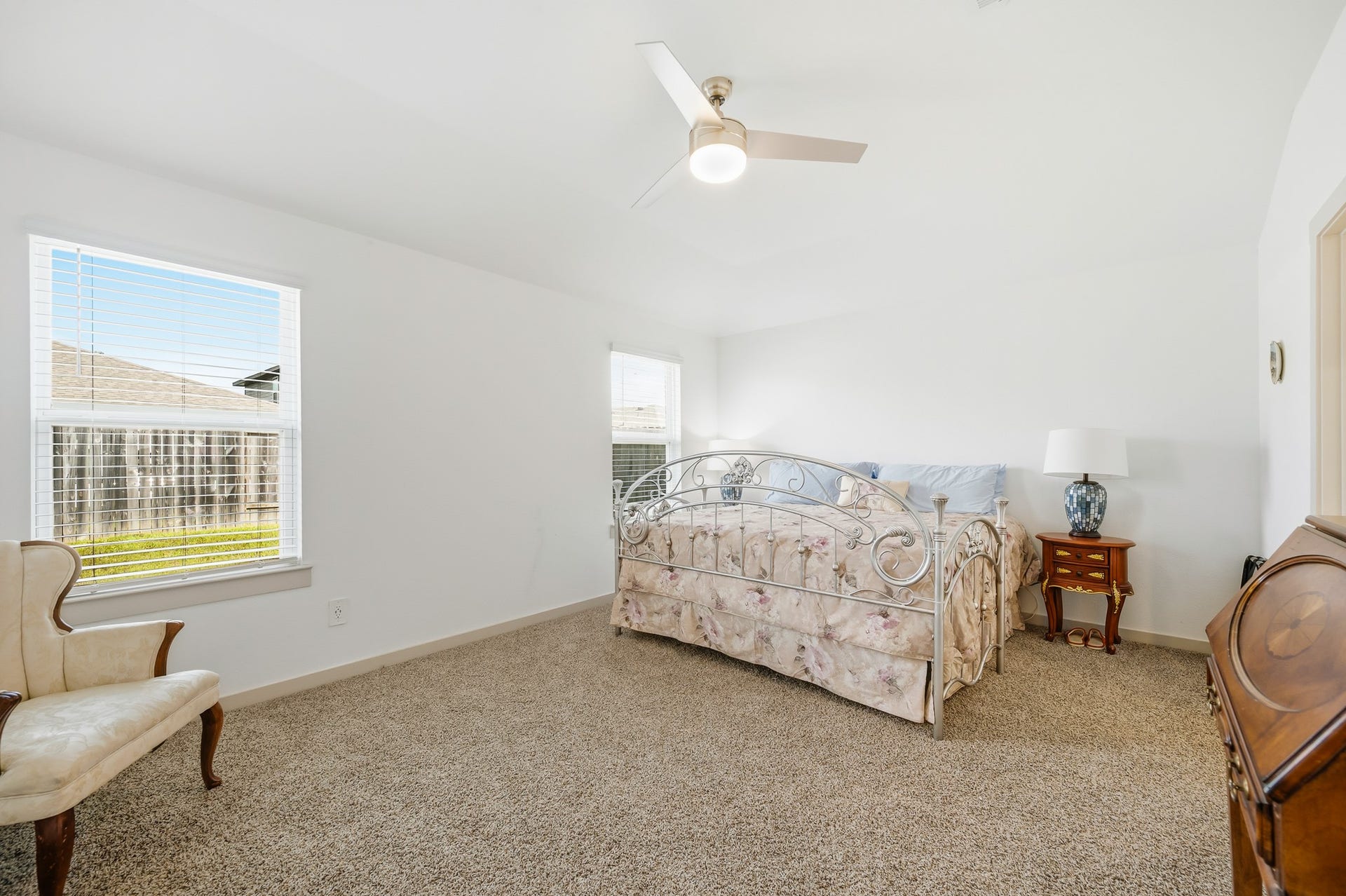 7002 Fleetwood Crescent Way, Richmond, TX 77407 This bright bedroom features neutral walls and carpet, a ceiling fan, and ample natural light from two windows. It includes a decorative metal bed and classic furnishings, creating a cozy and inviting atmosphere.