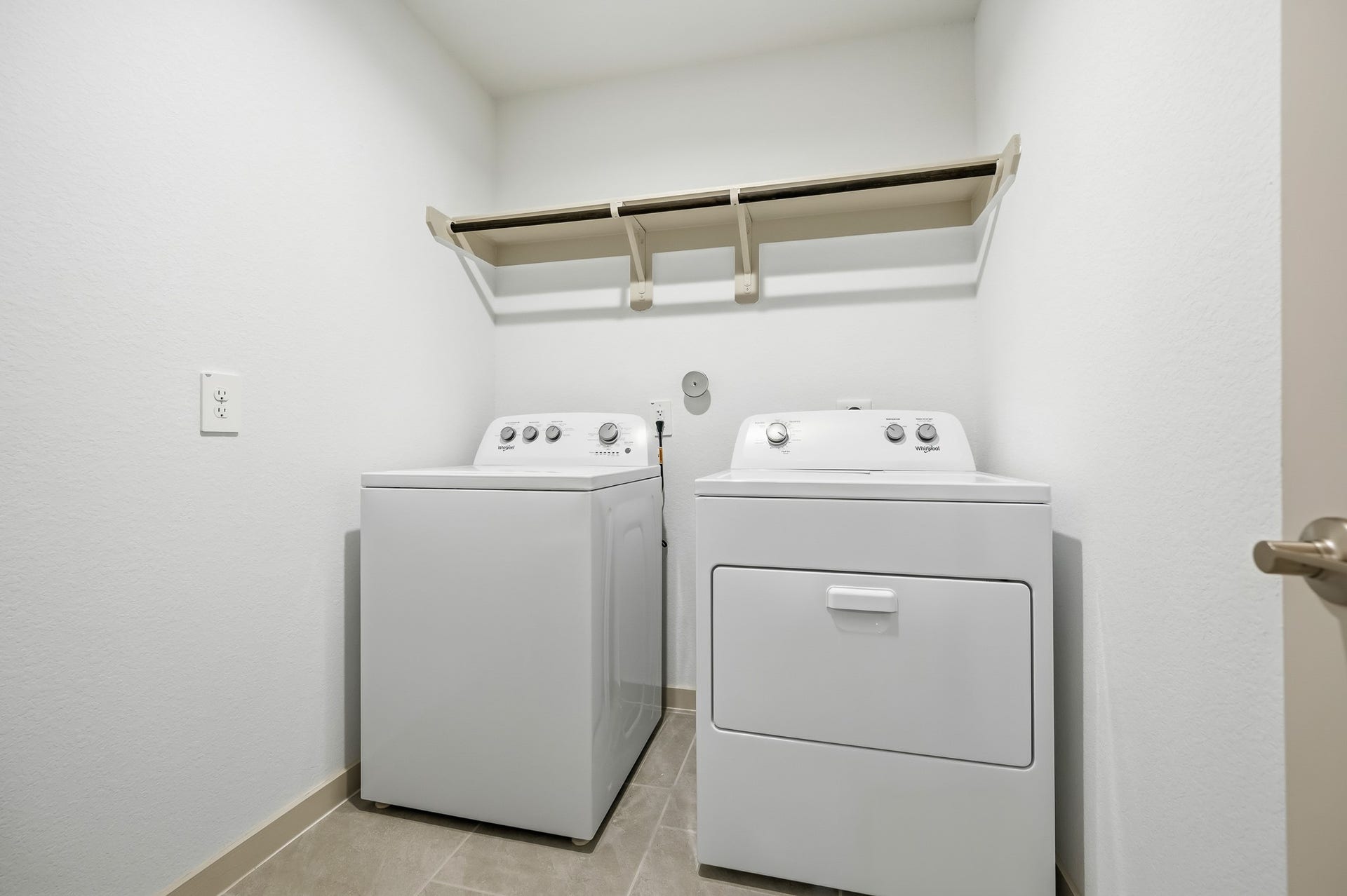 7002 Fleetwood Crescent Way, Richmond, TX 77407 This photo shows a compact laundry room with a washer and dryer set, neutral walls, tile flooring, and a shelf for storage.