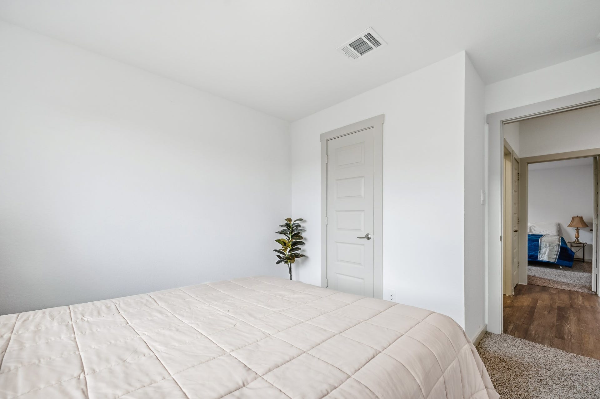 7002 Fleetwood Crescent Way, Richmond, TX 77407 This photo showcases a bright, minimalist bedroom with neutral walls and carpet. It features a neatly made bed, a small indoor plant, and a door leading to another room, creating an open feel.