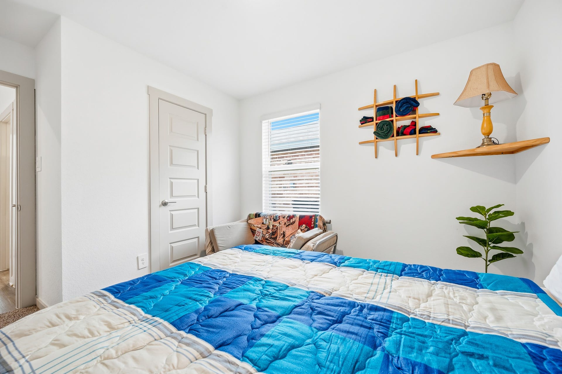 7002 Fleetwood Crescent Way, Richmond, TX 77407 Cozy bedroom featuring a bright window, simple decor, and a colorful bedspread. The room includes a small shelf with a lamp and a decorative plant, creating a welcoming and functional space.