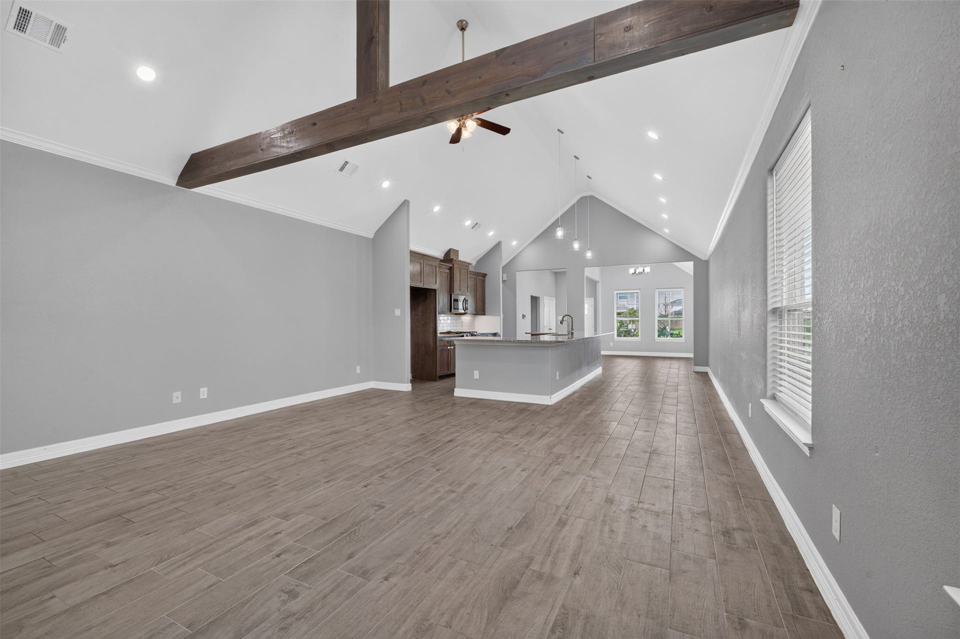 Spacious open-concept living area with a vaulted ceiling and exposed beam, featuring elegant wood-look tile flooring and abundant natural light from large windows.