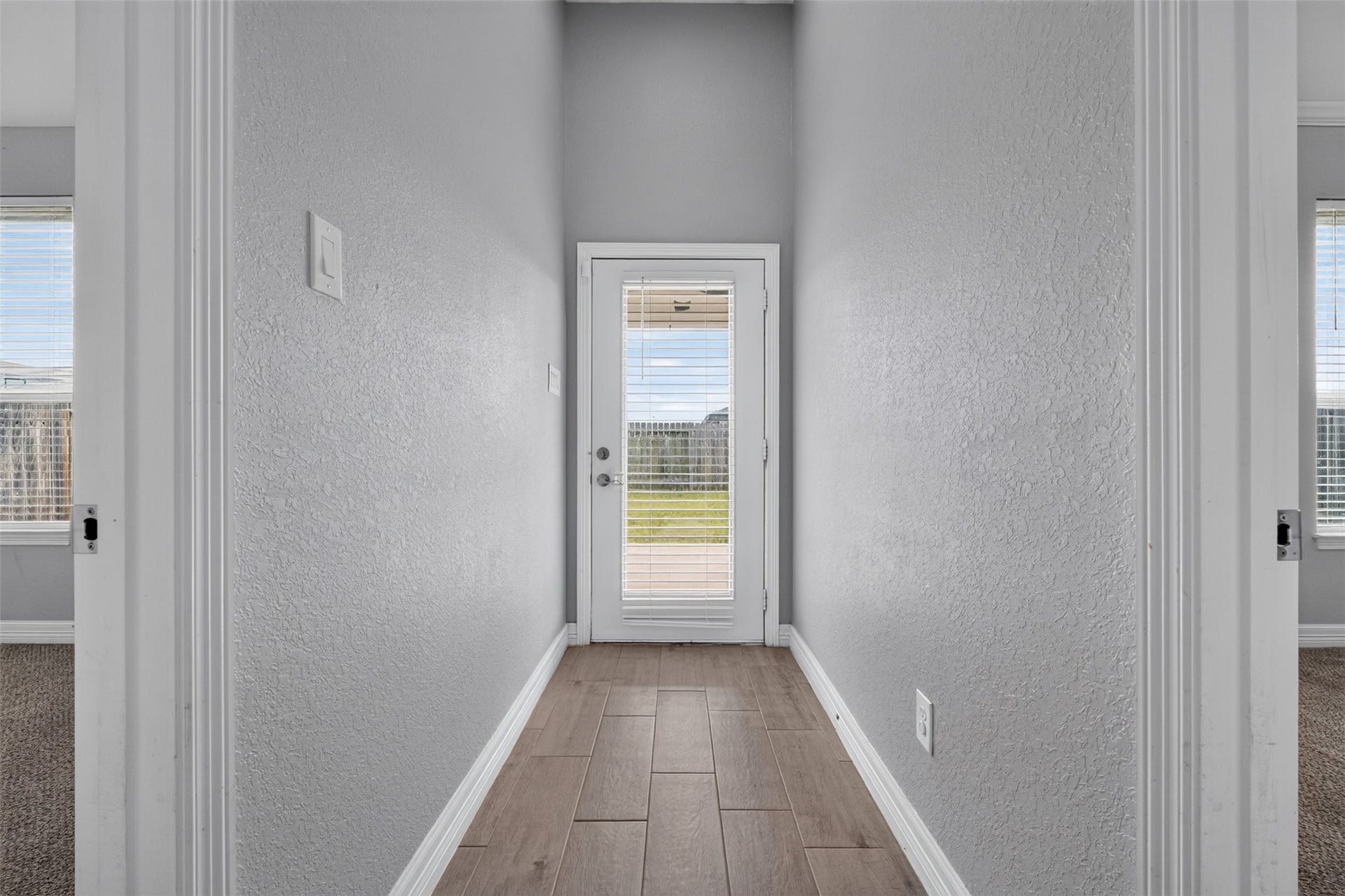 Bright hallway featuring sleek tile flooring and a glass door leading to a lush backyard, flanked by carpeted rooms on each side.