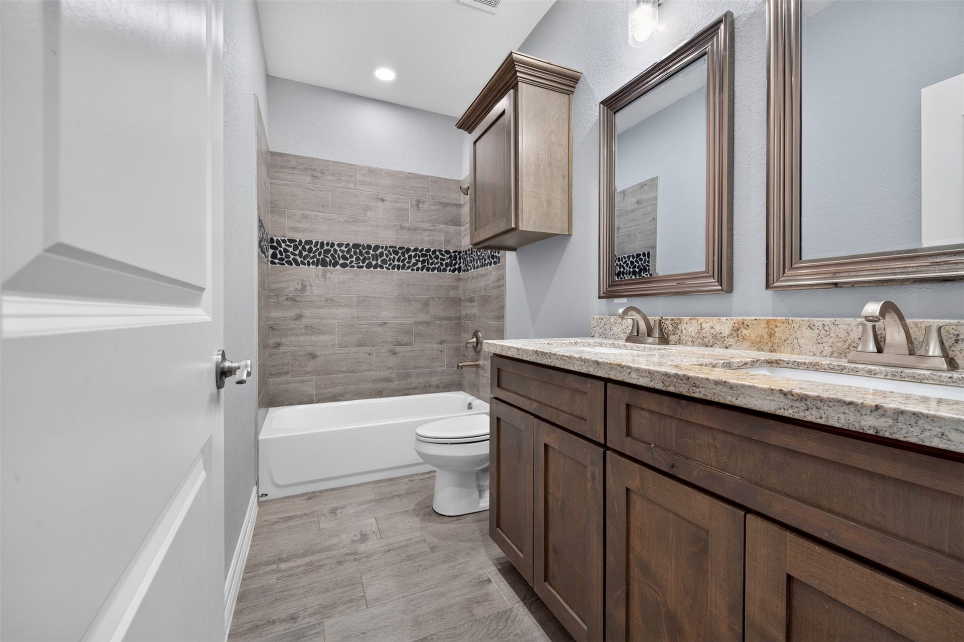 Elegant bathroom featuring dual sinks with granite countertops, rich wooden cabinetry, and a stylish tiled shower with a pebble accent strip.