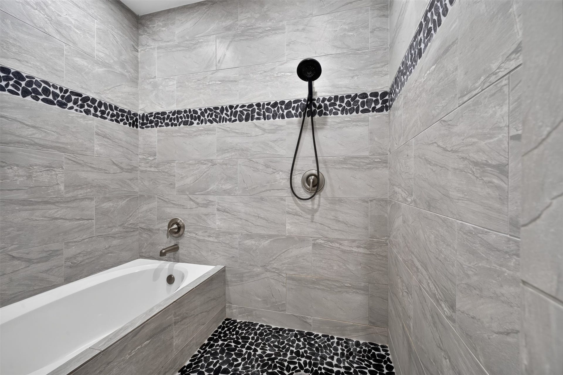 Elegant bathroom featuring a luxurious tub, sleek gray tiles with a stylish pebble accent strip, and a modern handheld showerhead for a spa-like experience.
