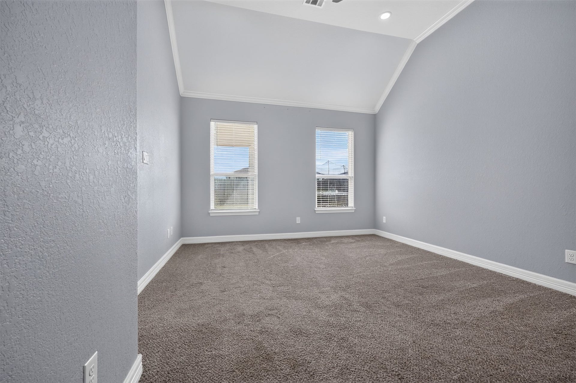 Spacious room with high vaulted ceilings, soft gray walls, and plush carpeting, featuring two large windows that invite natural light.