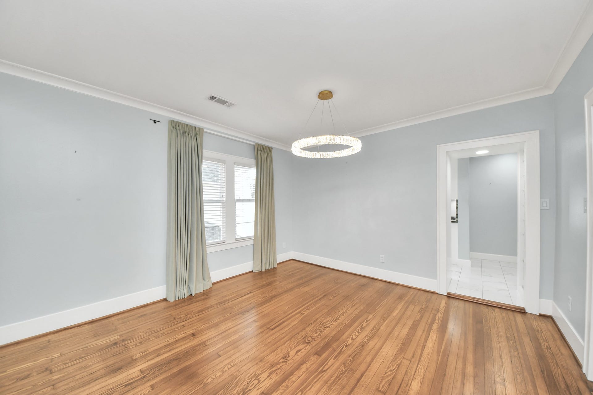 909 Fugate Street, Houston, TX 77009 Elegant room featuring polished hardwood floors, soft gray walls, and a contemporary chandelier. Natural light pours in through large windows with flowing curtains, creating a warm and inviting atmosphere.