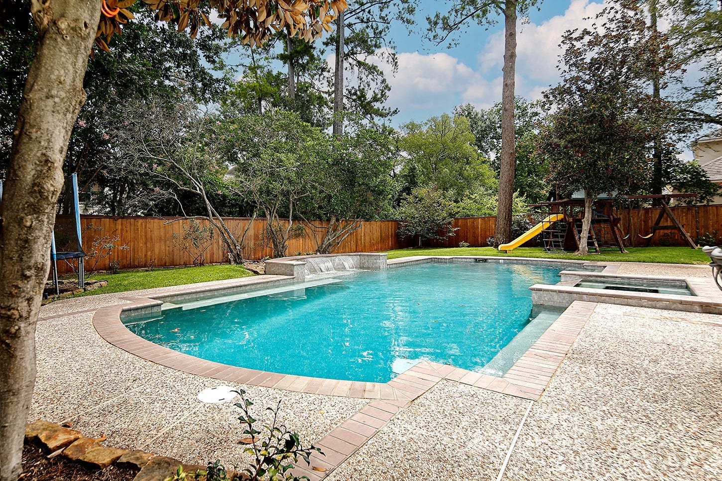 10 Snow Pond Place, The Woodlands, TX 77382 This exceptional backyard features a sprawling pool complete with recessed seating and water features, alongside ample space for a trampoline and play area.