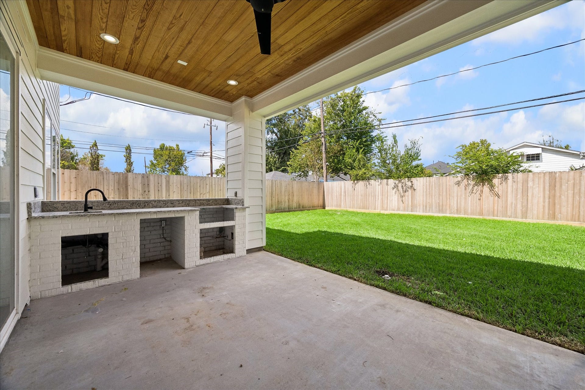 1726 Chantilly Lane, Houston, TX 77018 The covered patio features a wood ceiling, fan and a counter with an installed sink. The outdoor-kitchen base has open appliance spaces in this photo, with the fenced lawn just beyond.