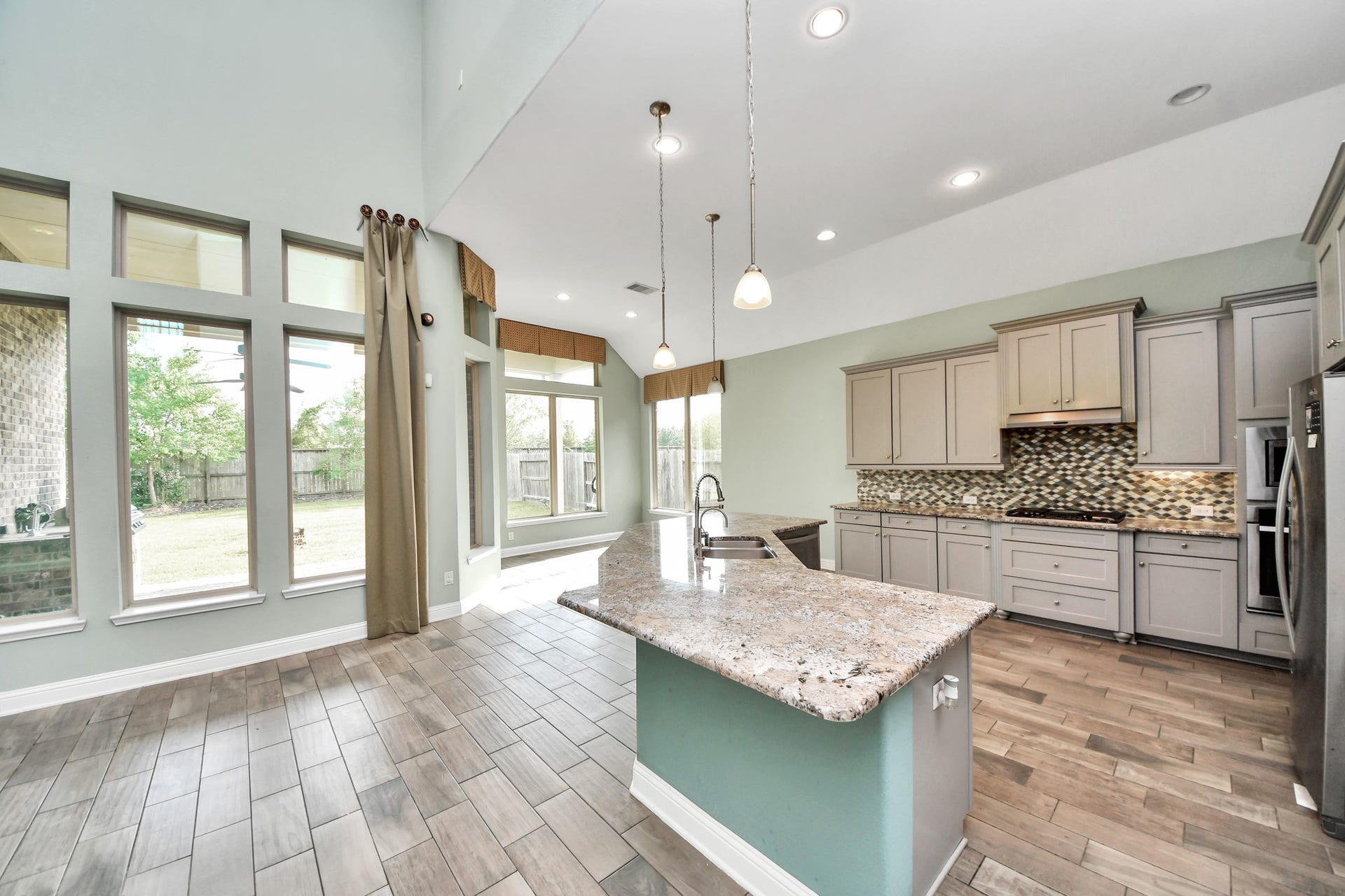 Bright open kitchen with granite island, abundant cabinetry, high ceilings, and walls of windows overlooking the backyard.