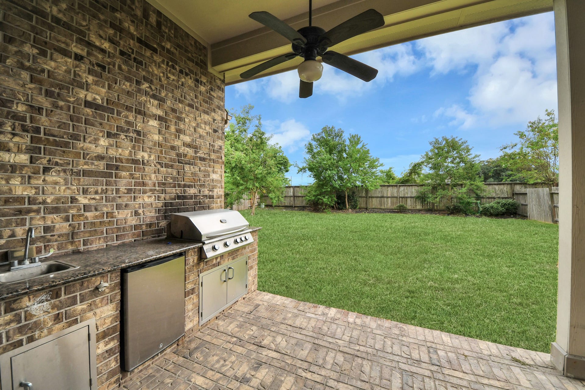 Enjoy outdoor living with a covered patio featuring a ceiling fan and built-in outdoor kitchen with grill, sink, countertop space, and storage, overlooking the spacious fenced backyard.