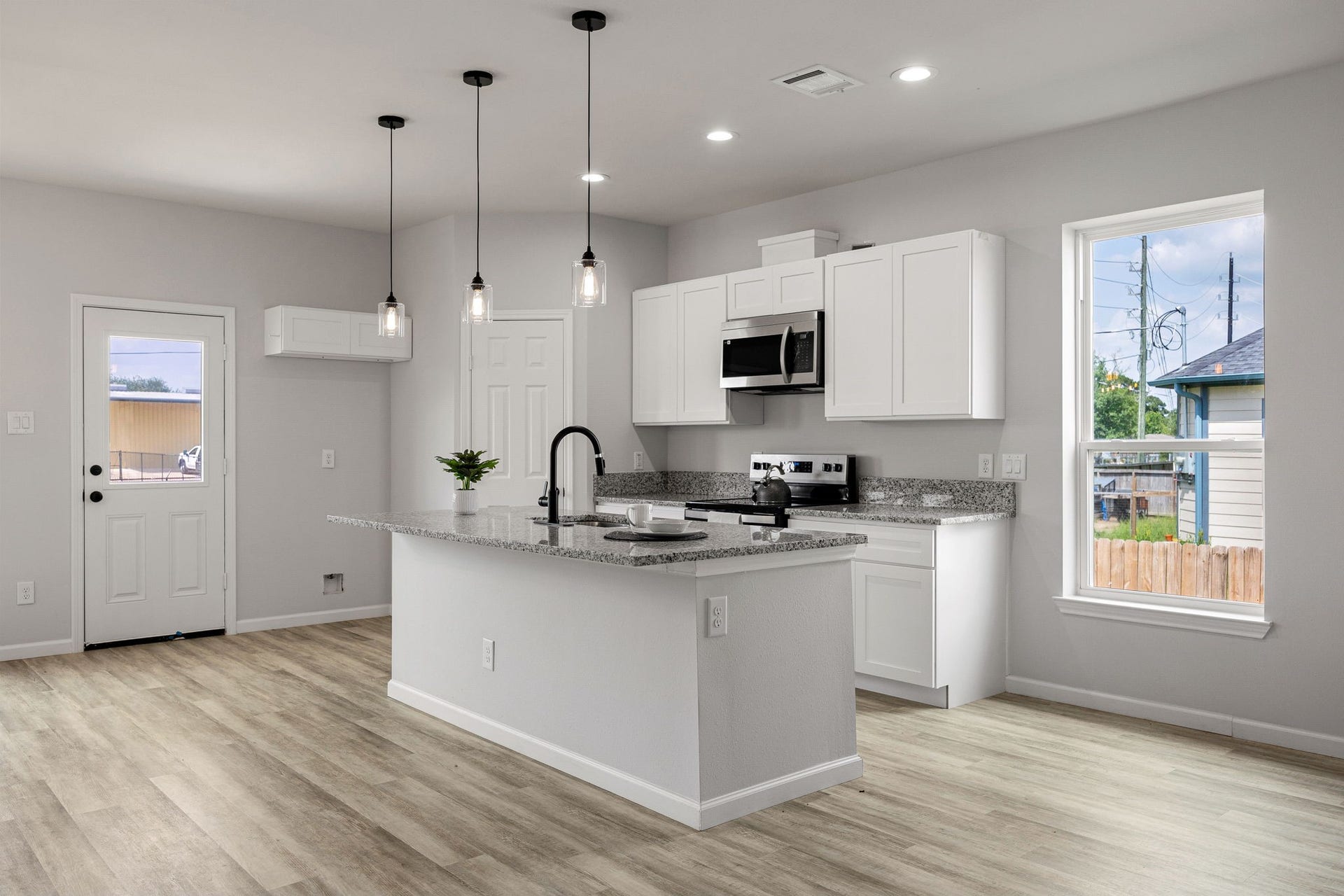 UNIT B- Kitchen, This modern kitchen features sleek white cabinetry, stainless steel appliances, and a granite countertop island with pendant lighting. The space is bright and airy, with light wood flooring and ample natural light from large windows.