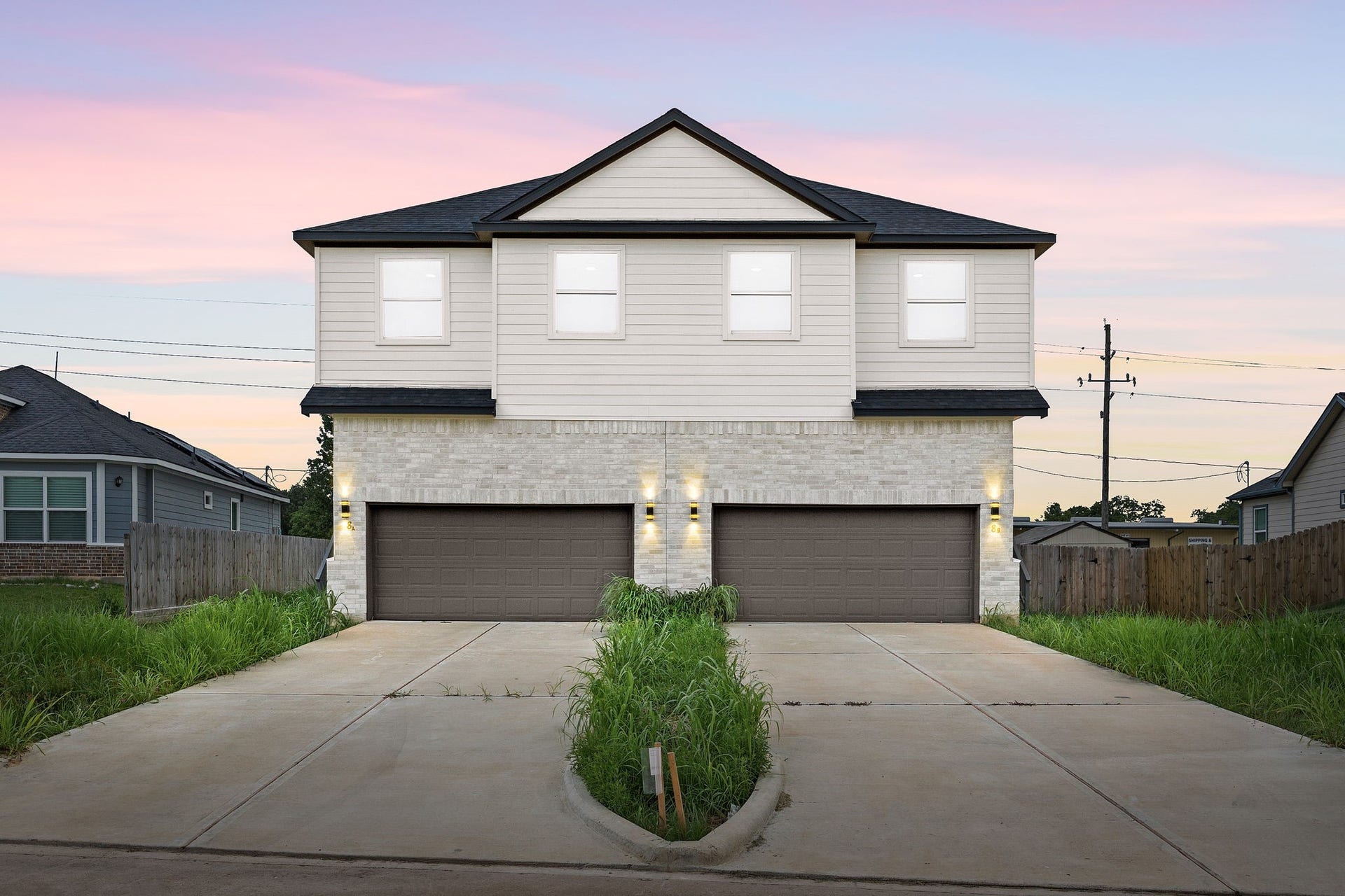 This is a modern duplex with a two-car garage, featuring a light brick and siding exterior. It has a spacious driveway and is set against a picturesque sunset sky.