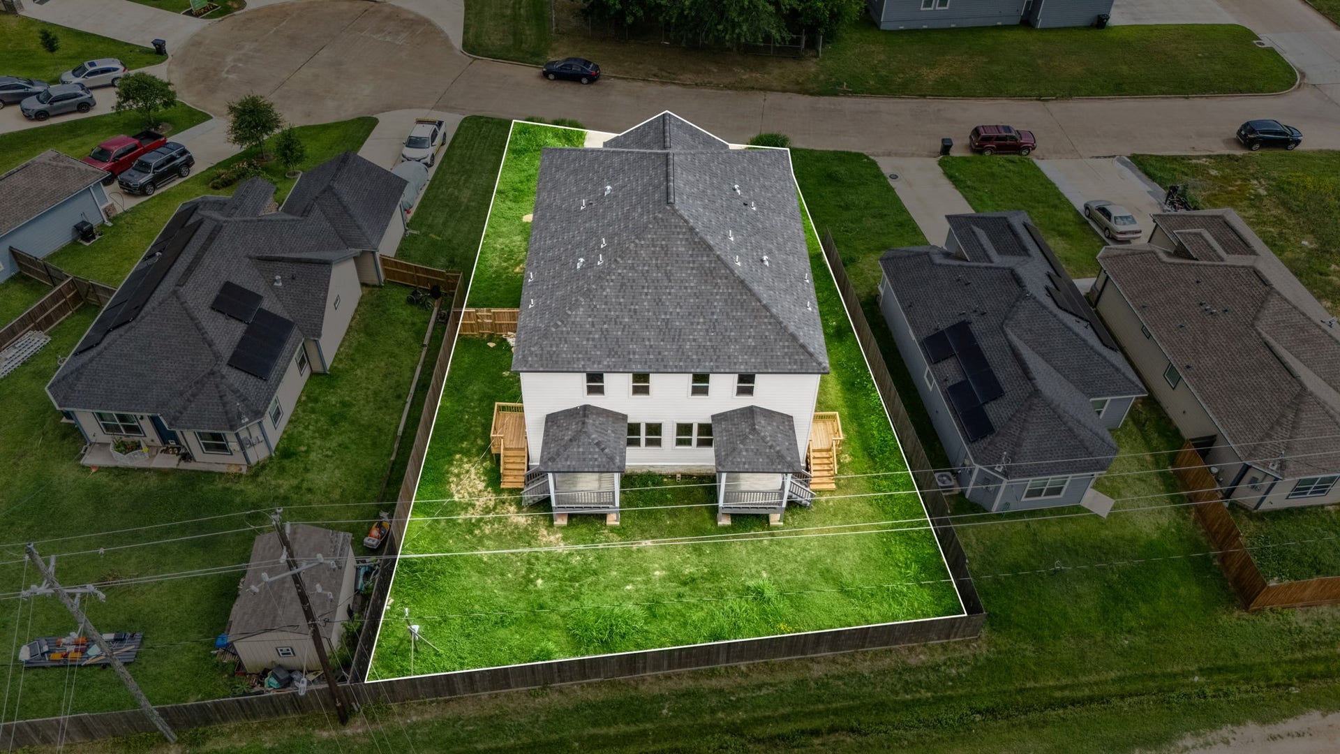 This aerial photo shows a two-story house with a gray roof and white exterior, set on a spacious fenced lot with a well-maintained lawn. The property is surrounded by neighboring homes and is located on a quiet street, offering a blend of community and privacy.