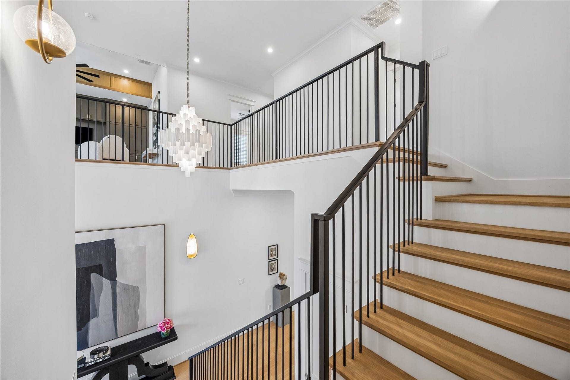 White oak stair treads and streamlined black iron railings create a striking architectural transition between levels. The open stairwell overlooks the two story foyer, allowing natural light and the statement chandelier to carry through both floors.