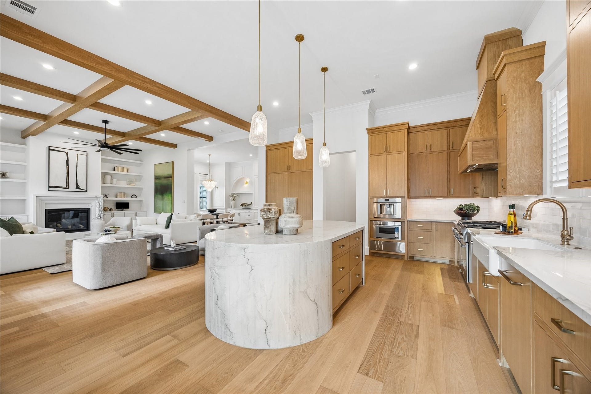 Oversized waterfall island anchors the kitchen while preserving an open sightline into the living area. Natural wood cabinetry, wide plank flooring and ceiling beams create a warm, cohesive palette throughout the main living spaces.