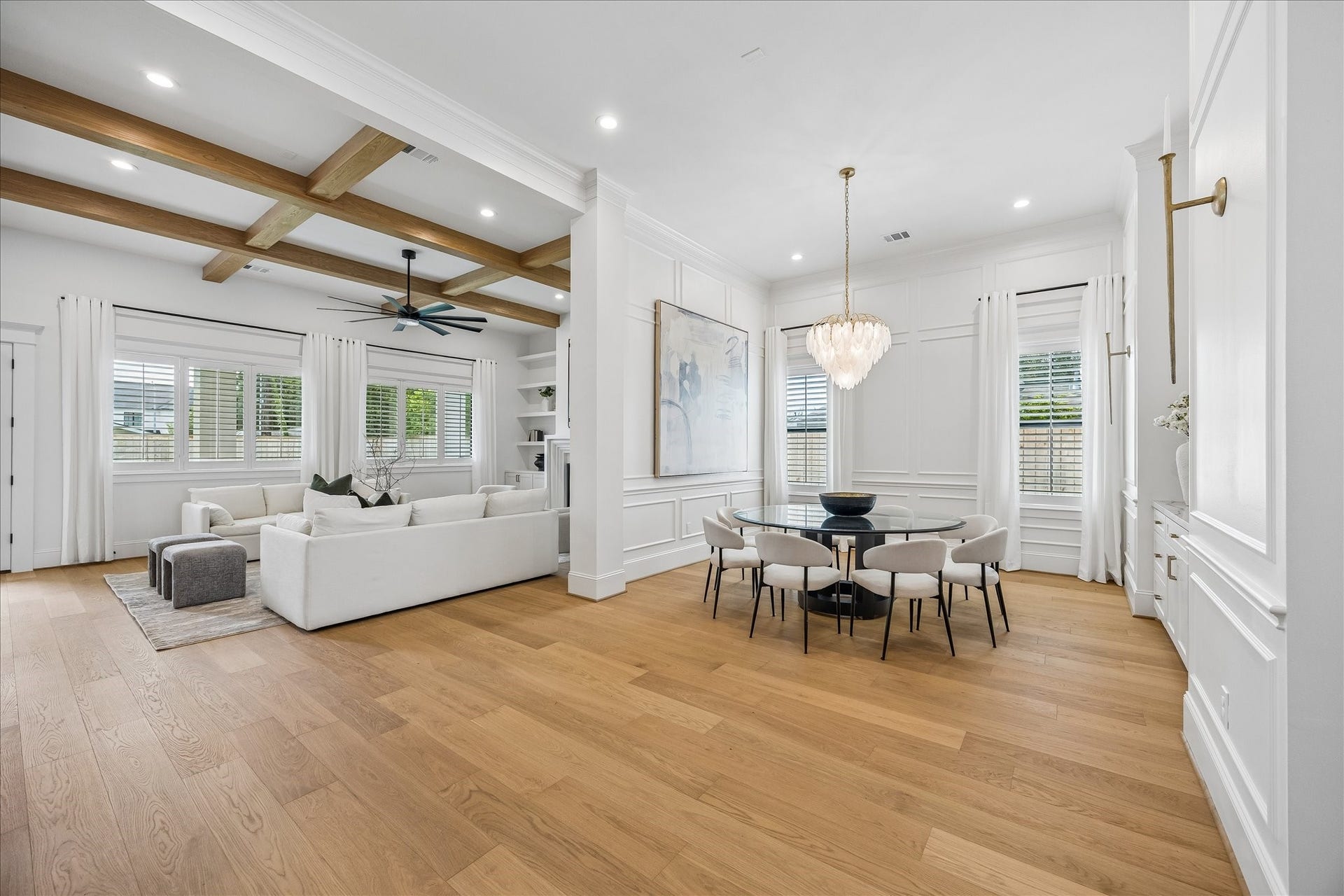 Open sightlines connect the formal dining area with the main living space, while wide plank hardwoods create a seamless flow between rooms. Detailed wall molding and a coffered ceiling with natural wood beams bring architectural character to the open layout.