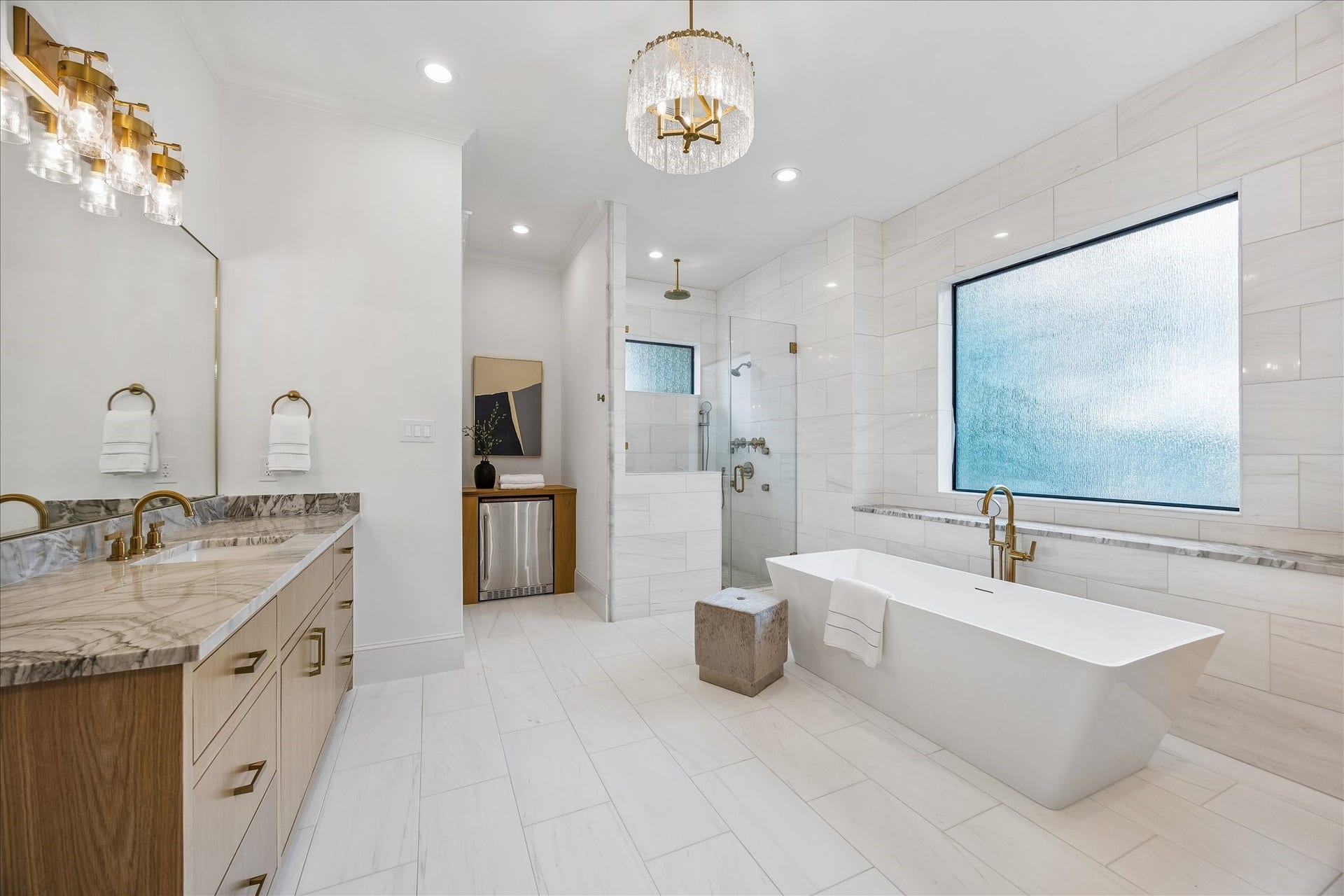 Spa inspired primary bath pairs floor to ceiling tile with a freestanding soaking tub, oversized glass enclosed shower and warm brass fixtures. A large privacy window fills the space with natural light.