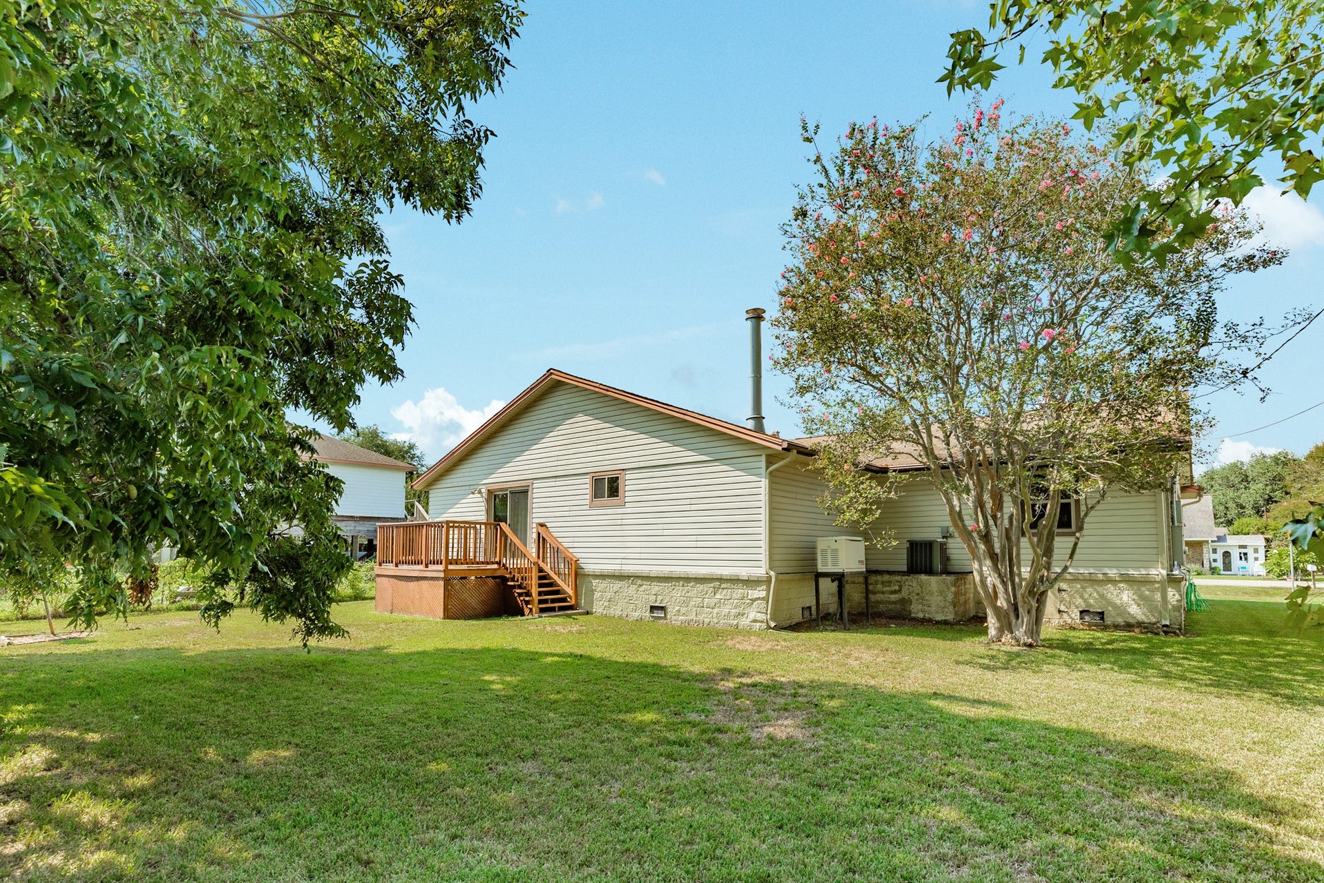 Spacious backyard provides abundant green space surrounded by mature trees, offering plenty of room for outdoor activities, gardening, or entertaining. A raised wood deck with stairs overlooks the yard and creates a convenient outdoor gathering space directly accessible from the home.