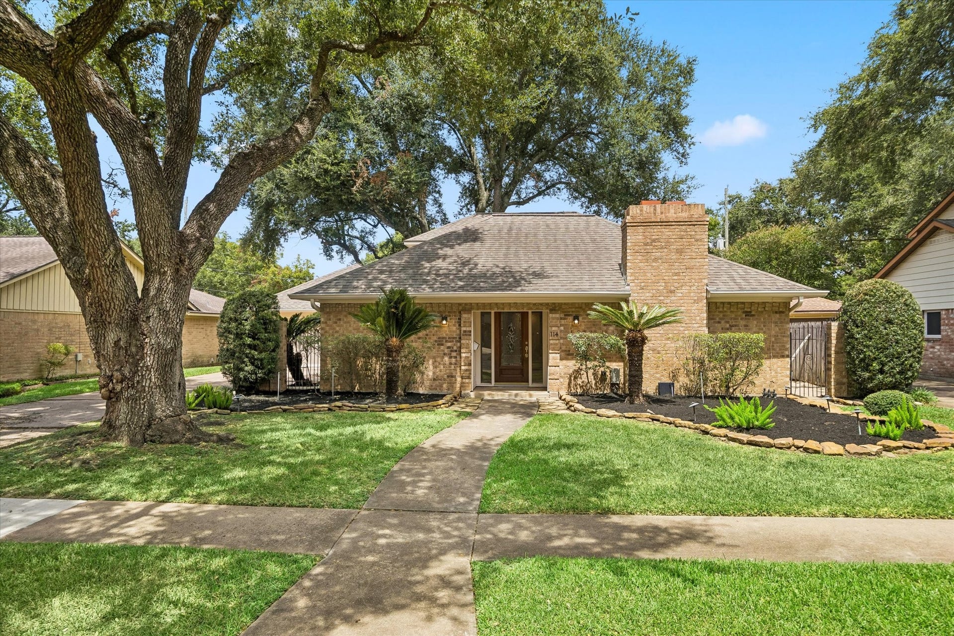 114 CAMELLIA STREET, Sugar Land, TX 77478 THE INVITING FRONT EXTERIOR SHOWCASES TIMELESS BRICK, A PROMINENT CHIMNEY, AND A WALKWAY FRAMED BY MATURE LANDSCAPING.