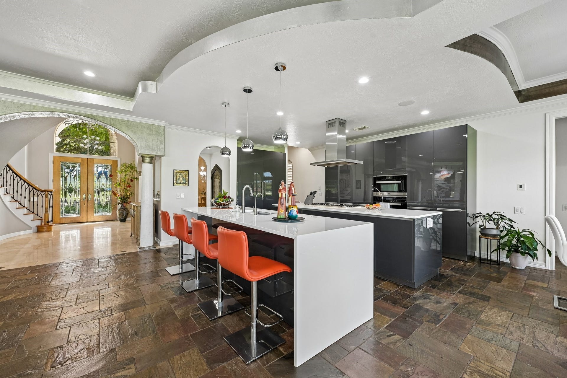 Contemporary kitchen featuring sleek dark cabinetry, integrated appliances, a statement vent hood, and sculptural ceiling details, with an open connection to the grand foyer