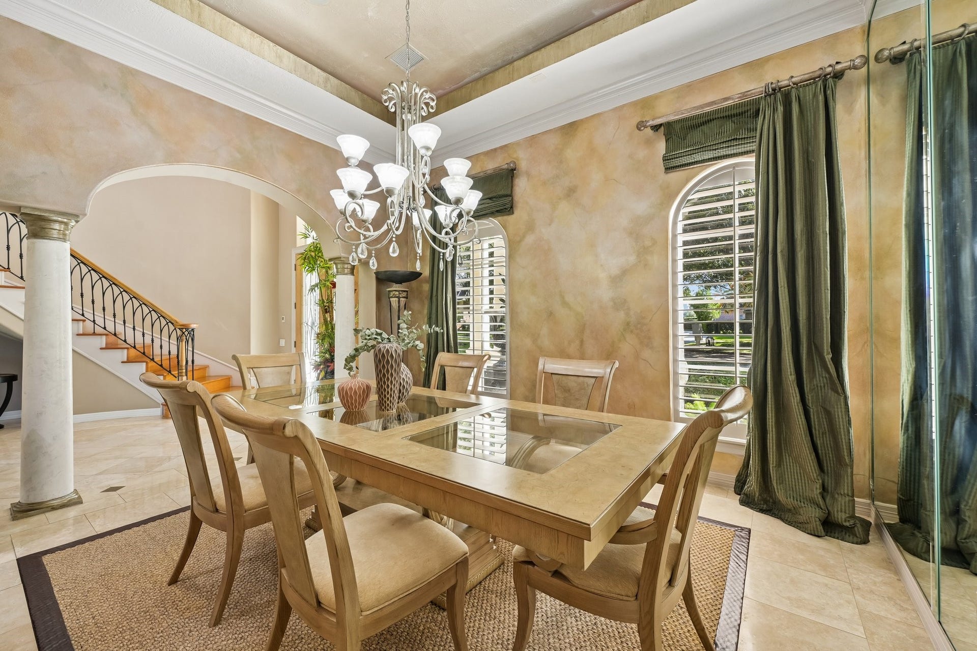 Elegant formal dining room featuring a statement chandelier, decorative ceiling detail, arched entryways, and expansive windows with plantation shutters
