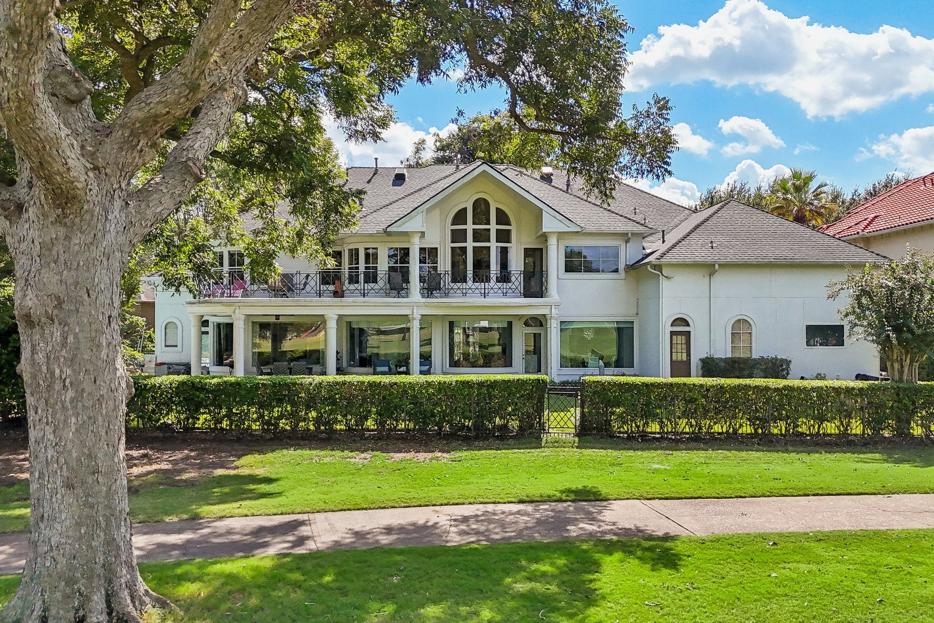 Rear exterior showcasing the home's expansive two-story design, covered patio, decorative wrought-iron railing, and mature tree framing the beautifully landscaped backyard