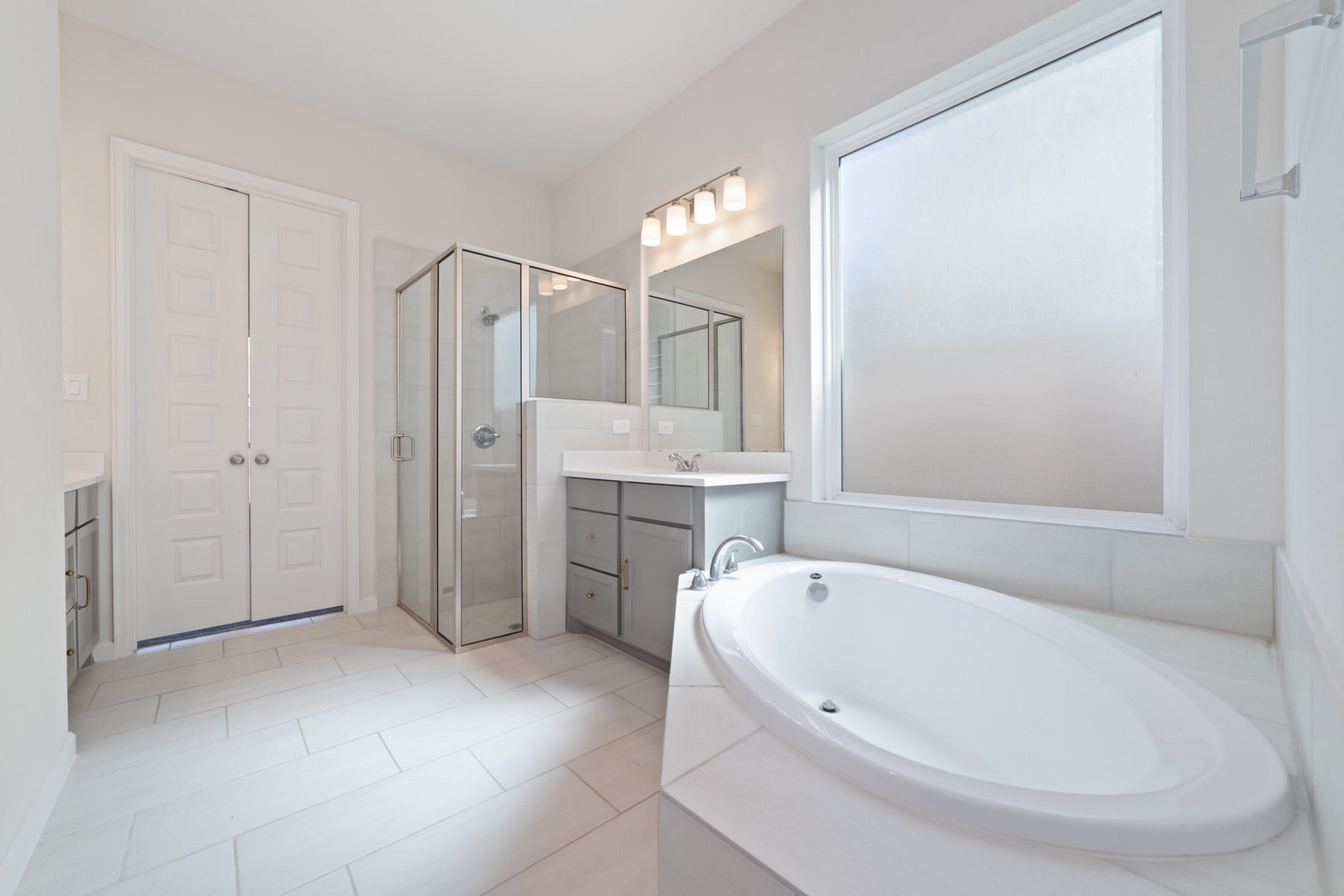 Bright and spacious bathroom featuring a luxurious soaking tub, sleek glass-enclosed shower, and modern dual vanities, all illuminated by natural light through a large frosted window.