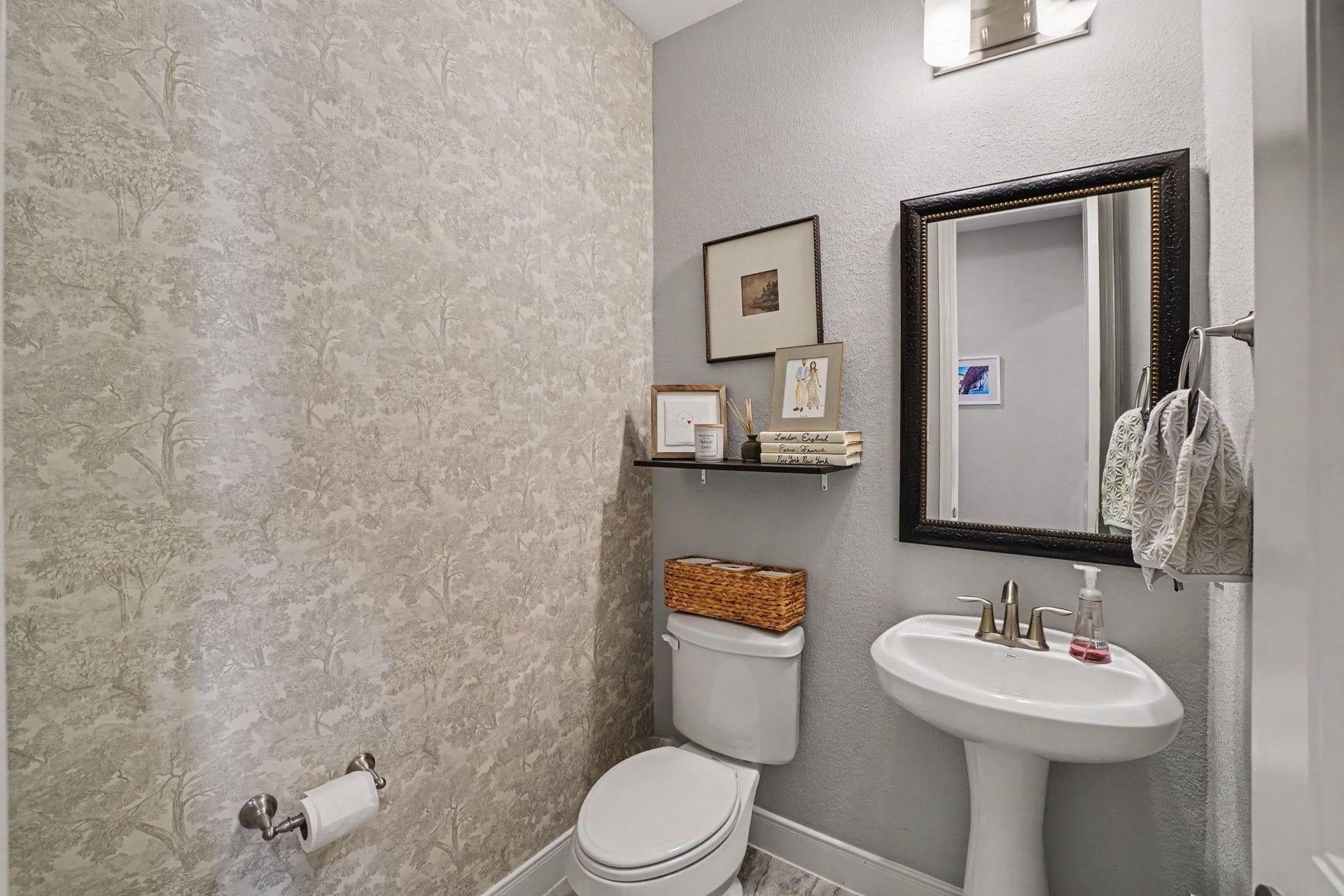26438 Morgan Creek Lane, Katy, TX 77494 This charming powder room offers a stylish and convenient space for visiting guests. One accent wall is dressed in a subtle, elegant tree-patterned wallpaper, adding personality and texture to the room. A classic white pedestal sink with brushed nickel hardware pairs with a dark-framed rectangular mirror and warm overhead lighting.