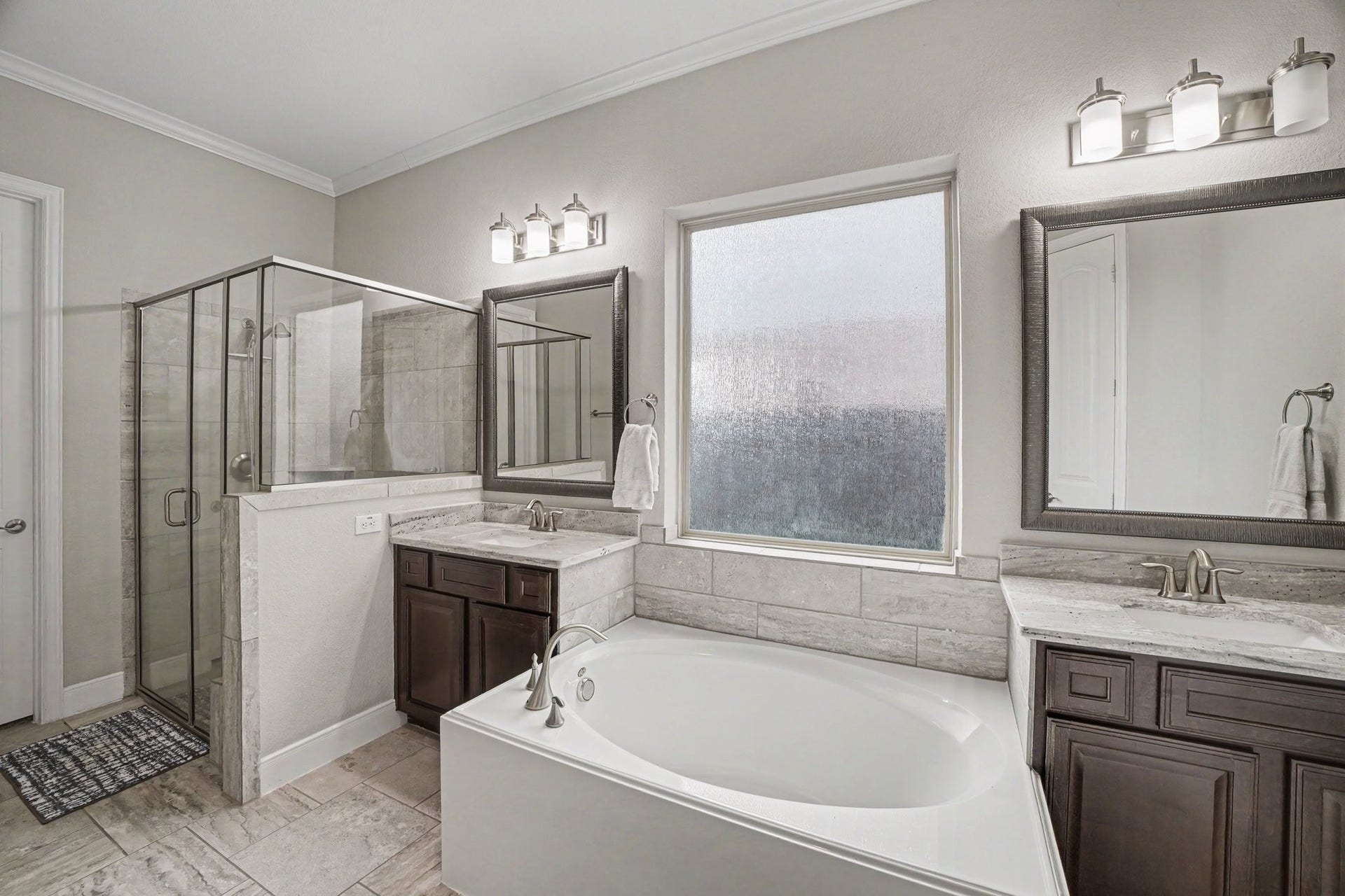 26438 Morgan Creek Lane, Katy, TX 77494 At the heart of the space lies a deep, oval soaking tub nestled beneath a large frosted privacy window, filling the room with soft natural light while maintaining complete seclusion. Flanking the tub are separate dual vanities featuring rich dark cabinetry, sleek granite countertops, brushed nickel hardware, framed mirrors, and contemporary three-light vanity fixtures.