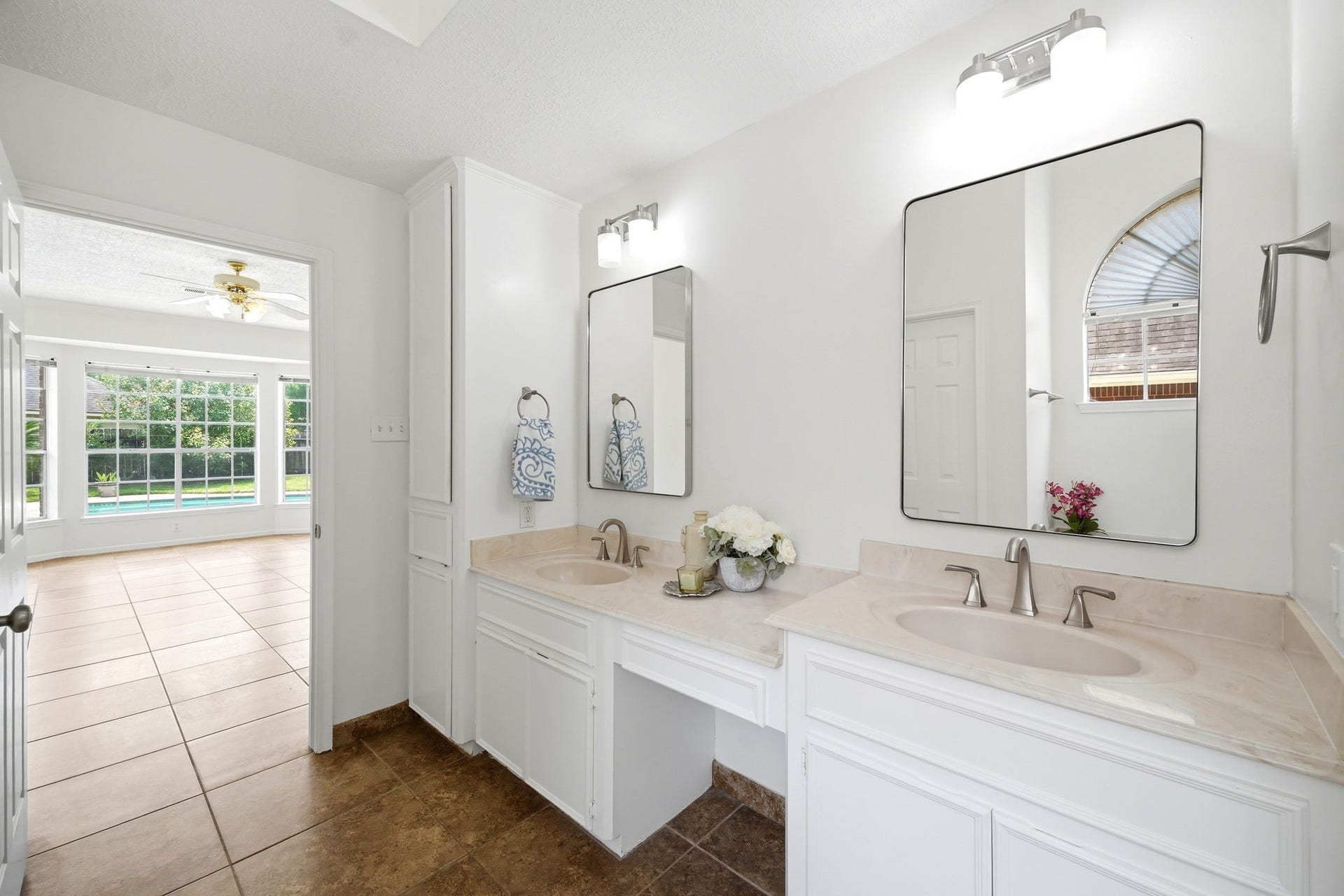 2216 Fairwater Park Drive, League City, TX 77573 Experience luxury in this bright, spacious bathroom with dual sinks, modern lighting, and expansive mirrors, leading into a sunlit room with tiled flooring and a large window showcasing a pool view.