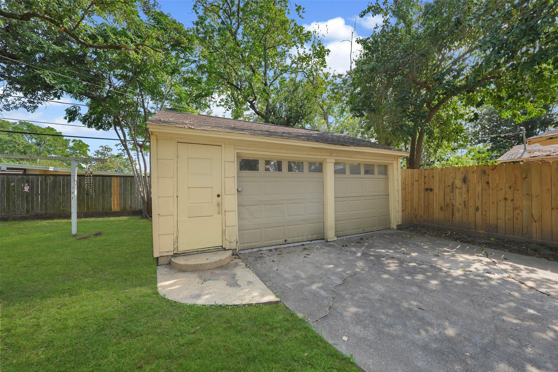 9519 Val Verde Street, Houston, TX 77063 This photo shows a two-car garage with a light yellow exterior, nestled in a fenced backyard. The surrounding area is spacious with green grass and mature trees, providing shade and privacy.