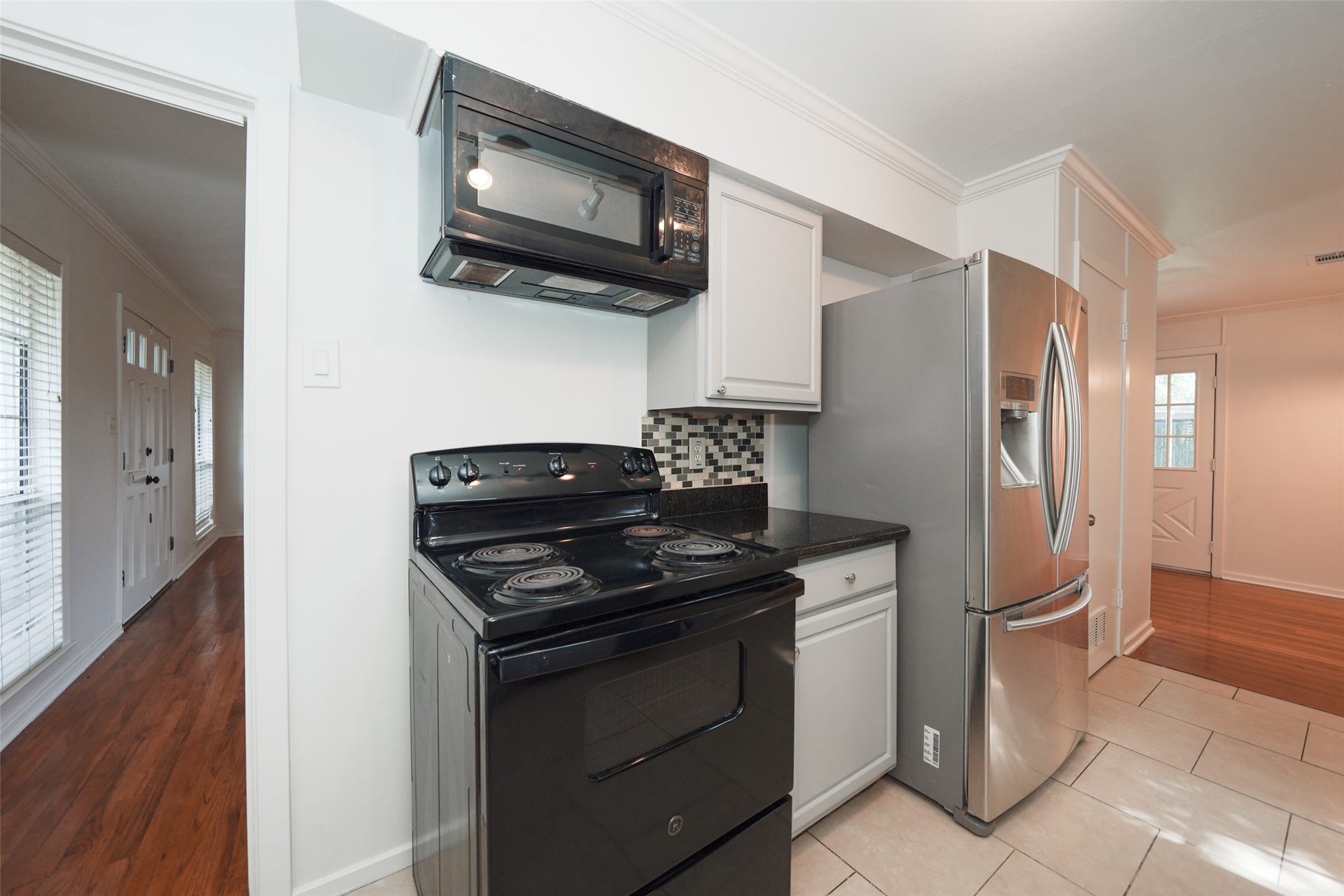 9519 Val Verde Street, Houston, TX 77063 This kitchen features a black stove and microwave, stainless steel refrigerator, and modern tile backsplash. It opens into a hallway with wood flooring and ample natural light from nearby windows.