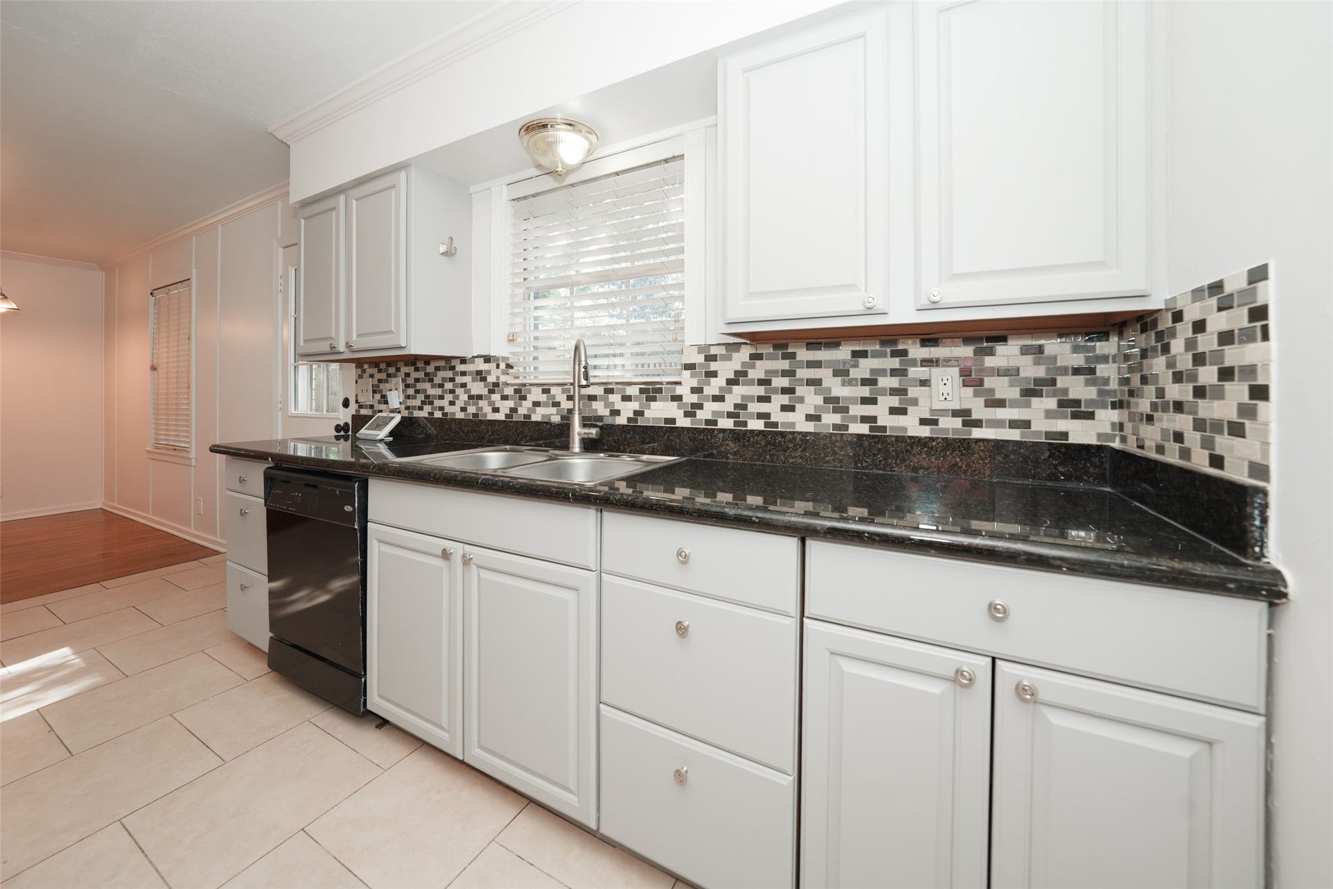 9519 Val Verde Street, Houston, TX 77063 This kitchen features modern white cabinetry, sleek black countertops, and a stylish mosaic tile backsplash. It includes a built-in dishwasher and a double sink beneath a window, allowing for plenty of natural light. The space seamlessly connects to the dining area with a combination of tile and wood flooring.