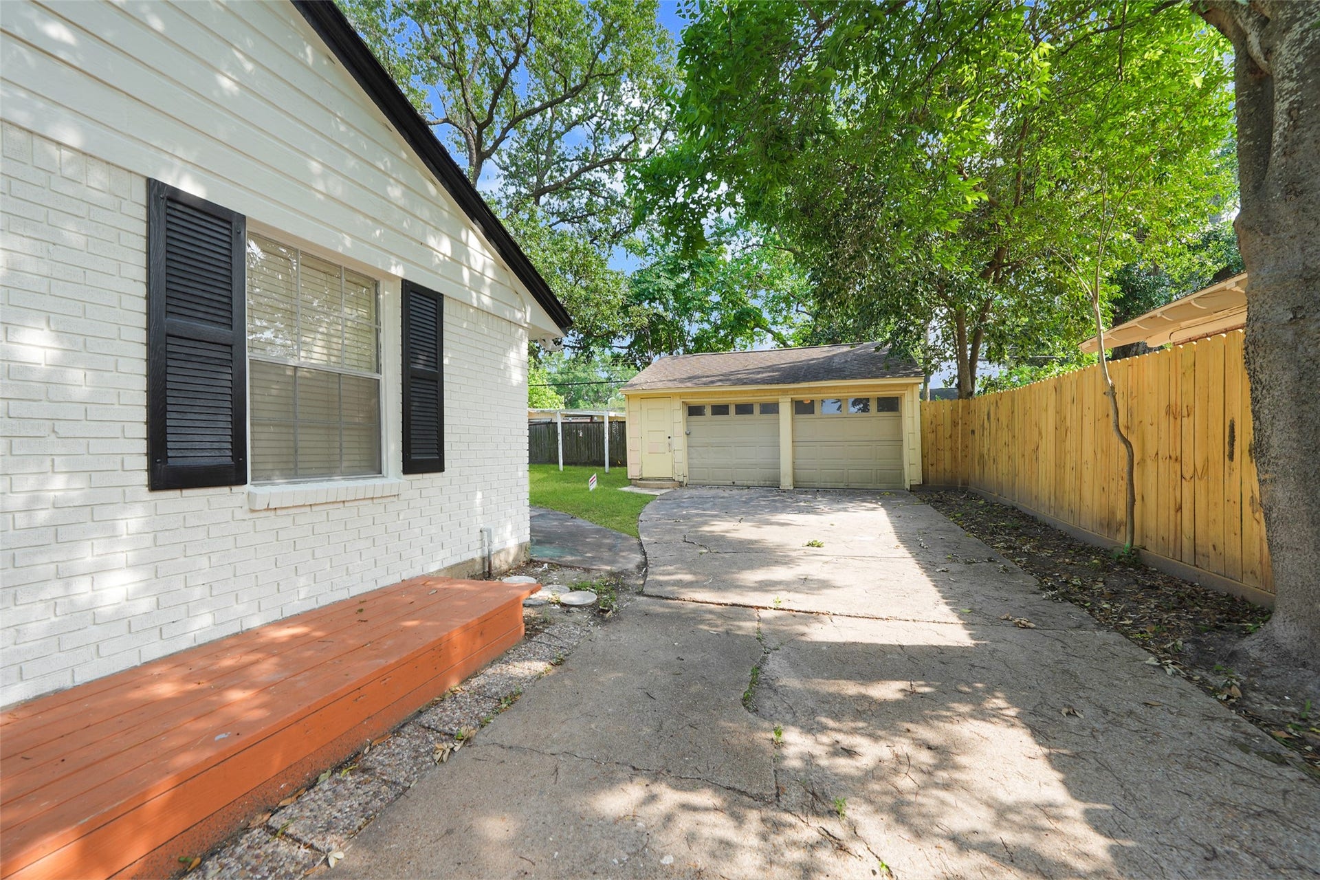9519 Val Verde Street, Houston, TX 77063 This photo shows a charming backyard view featuring a white brick house with black shutters, a wooden step, and a spacious driveway leading to a detached two-car garage. The area is partially shaded by mature trees and bordered by a wooden fence, offering a blend of privacy and natural beauty.