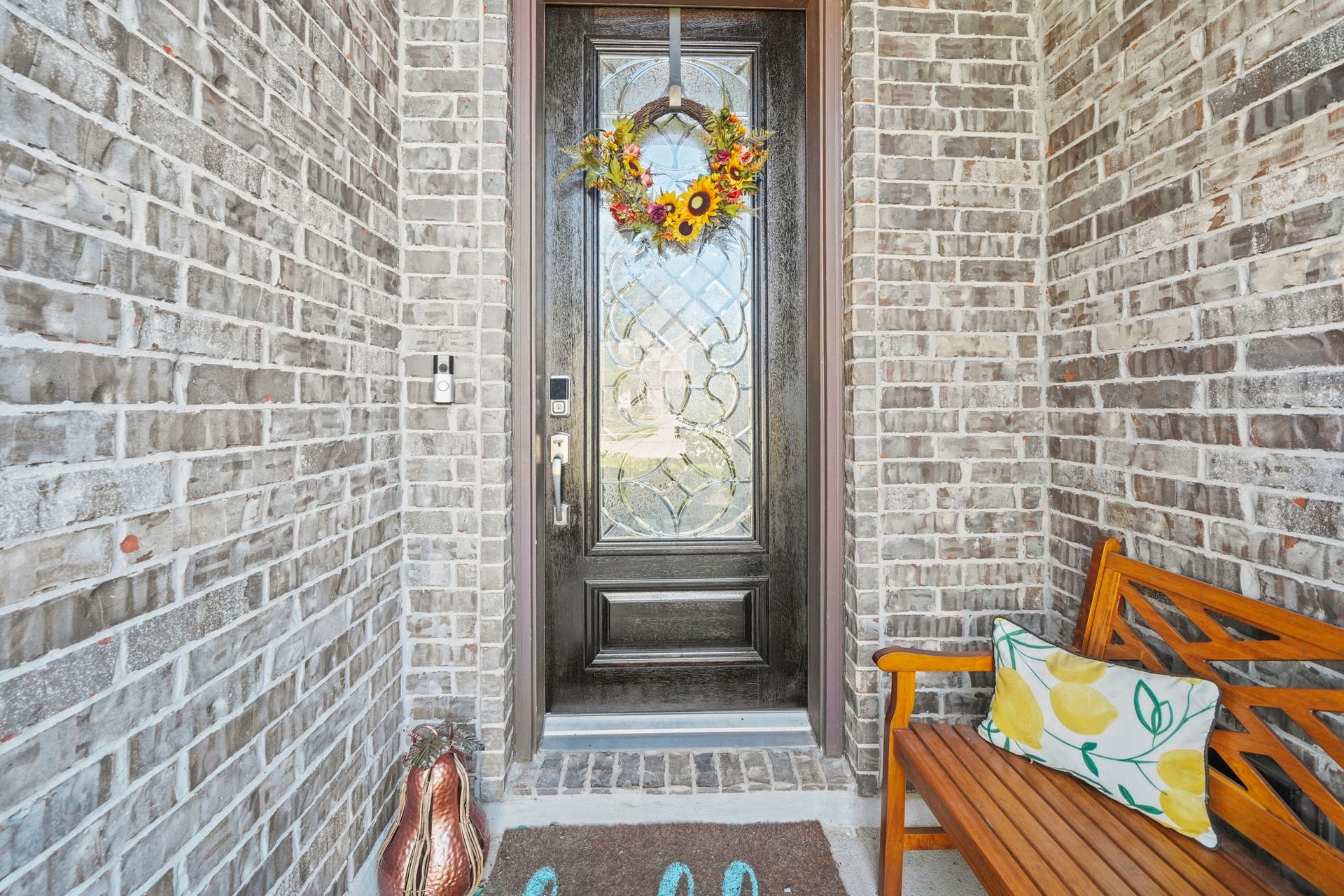 28107 Scenic Shore Lane, Fulshear, TX 77441 Charming brick entryway with a decorative glass front door, adorned with a vibrant floral wreath. Cozy bench seating adds a welcoming touch.