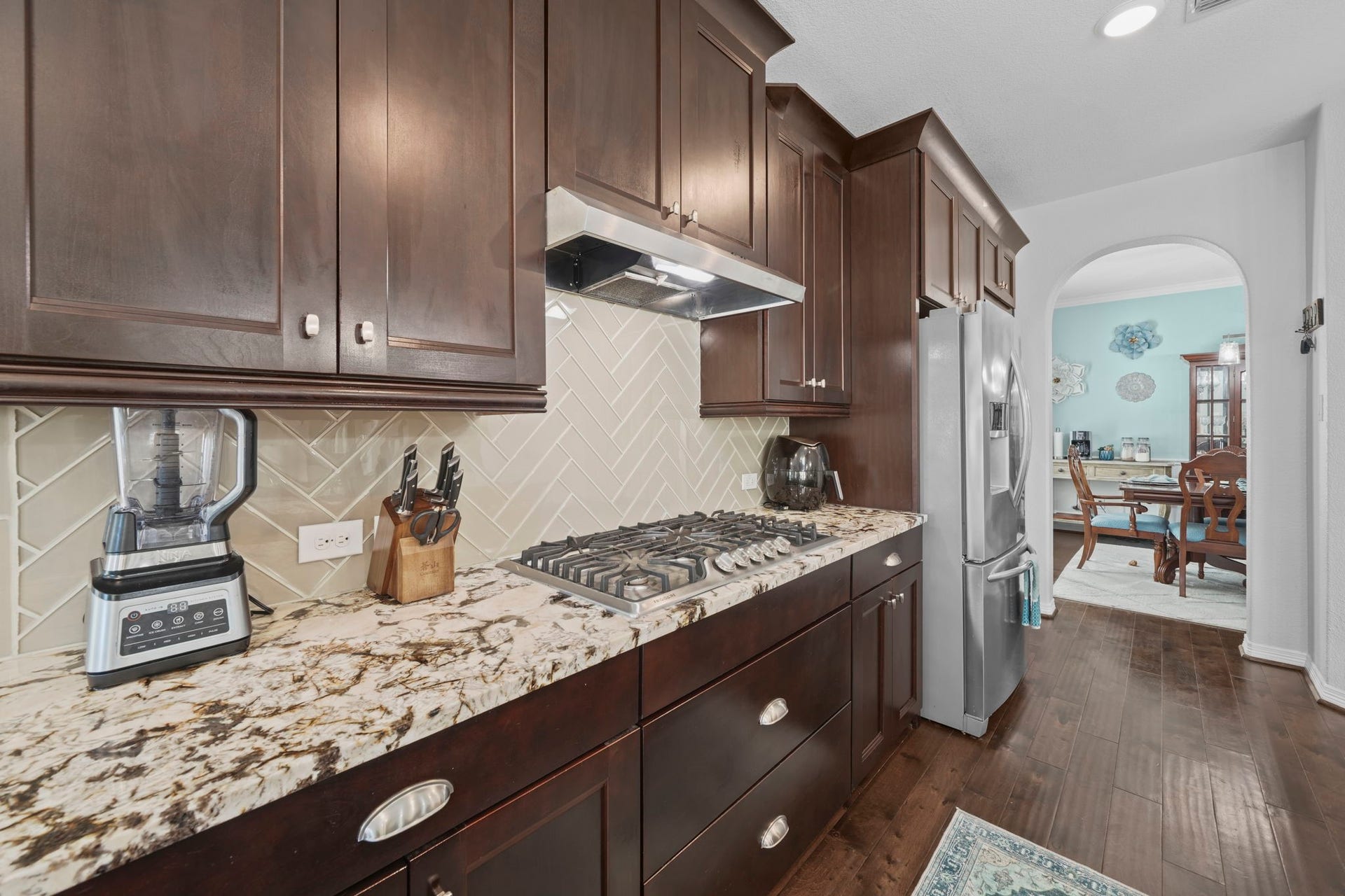 28107 Scenic Shore Lane, Fulshear, TX 77441 Elegant kitchen with rich dark wood cabinets, sleek stainless steel appliances, and a stylish herringbone tile backsplash. The granite countertops provide ample workspace, while the open archway leads to a cozy dining area.