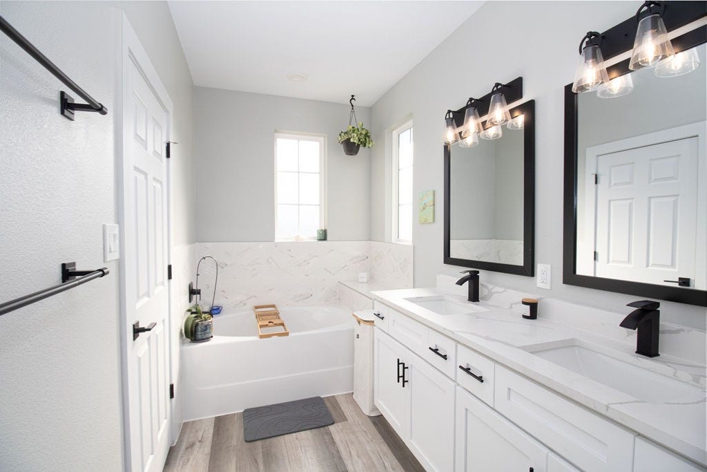 8415 Sunnyhill Street, #B, Houston, TX 77088 Relax and unwind in this beautifully appointed primary bathroom featuring a soaking tub, elegant marble-look tile accents, white cabinetry, stylish black fixtures, and natural light from two windows.