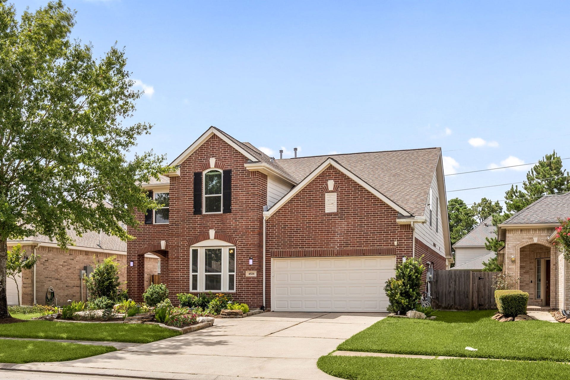 Timeless red brick exterior showcasing a grand gabled roofline, elegant shuttered windows, and a wide concrete driveway. Nestled on a beautifully landscaped lot with mature trees and a pristine green lawn.